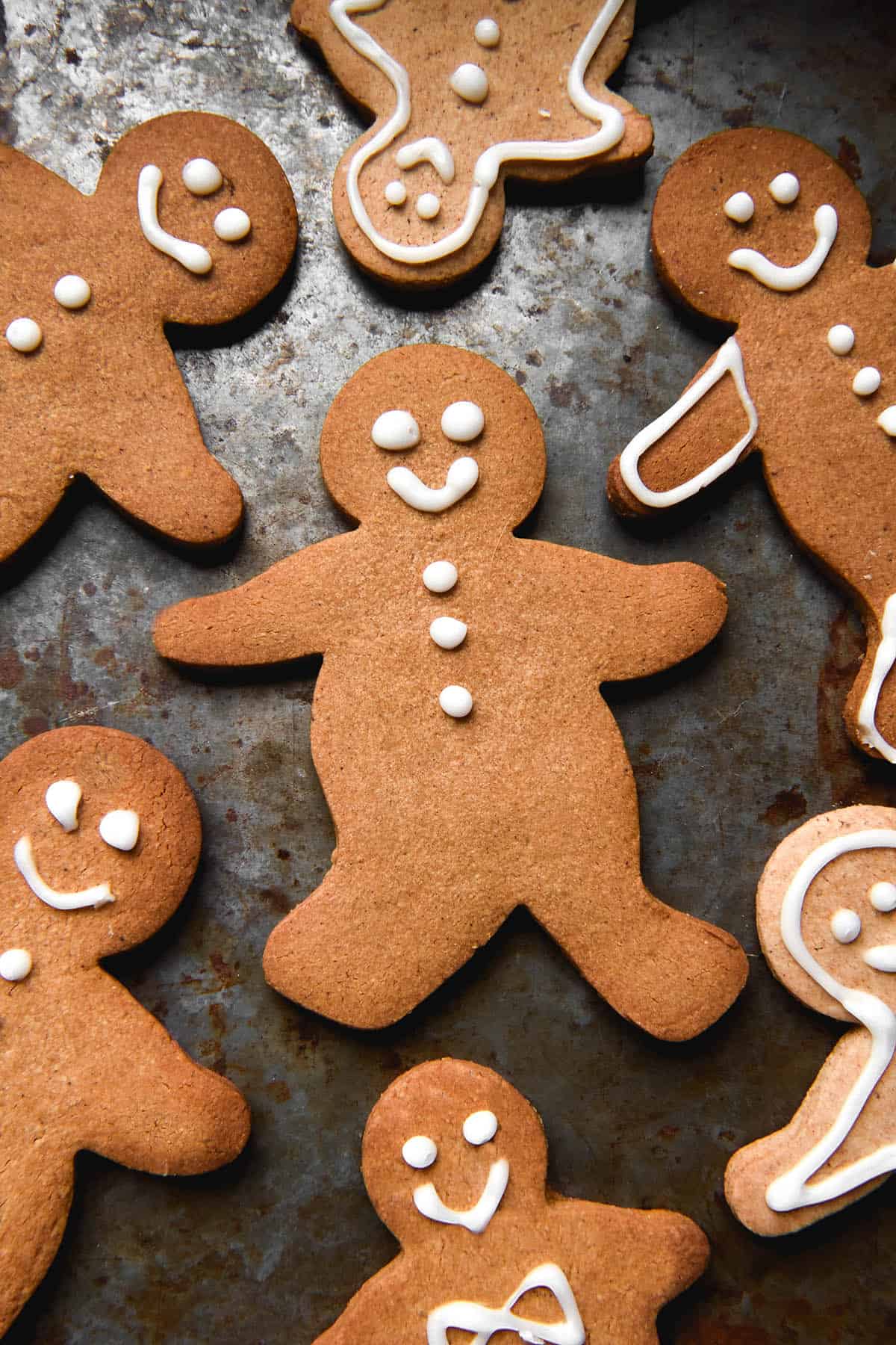 An aerial view of gluten free gingerbread people on a dark steel backdrop. The gingerbread are lightly decorated with white icing, giving them smiley faces and other light details. 