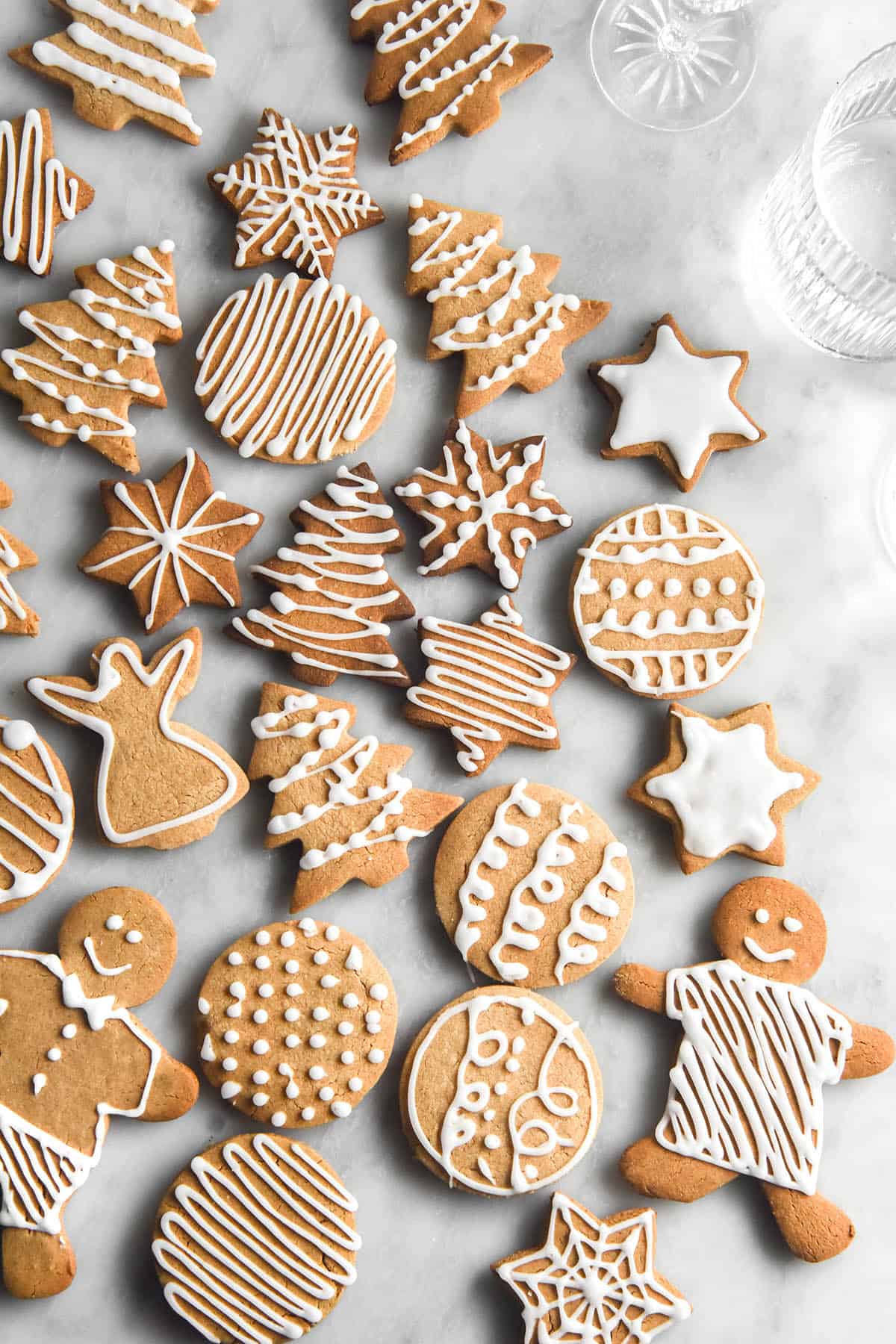 An aerial image of a group of variously shaped gluten free gingerbread atop a white marble table. The bauble, star, Christmas tree and gingerbread person shapes are all decorated with white icing sugar in different ways. 