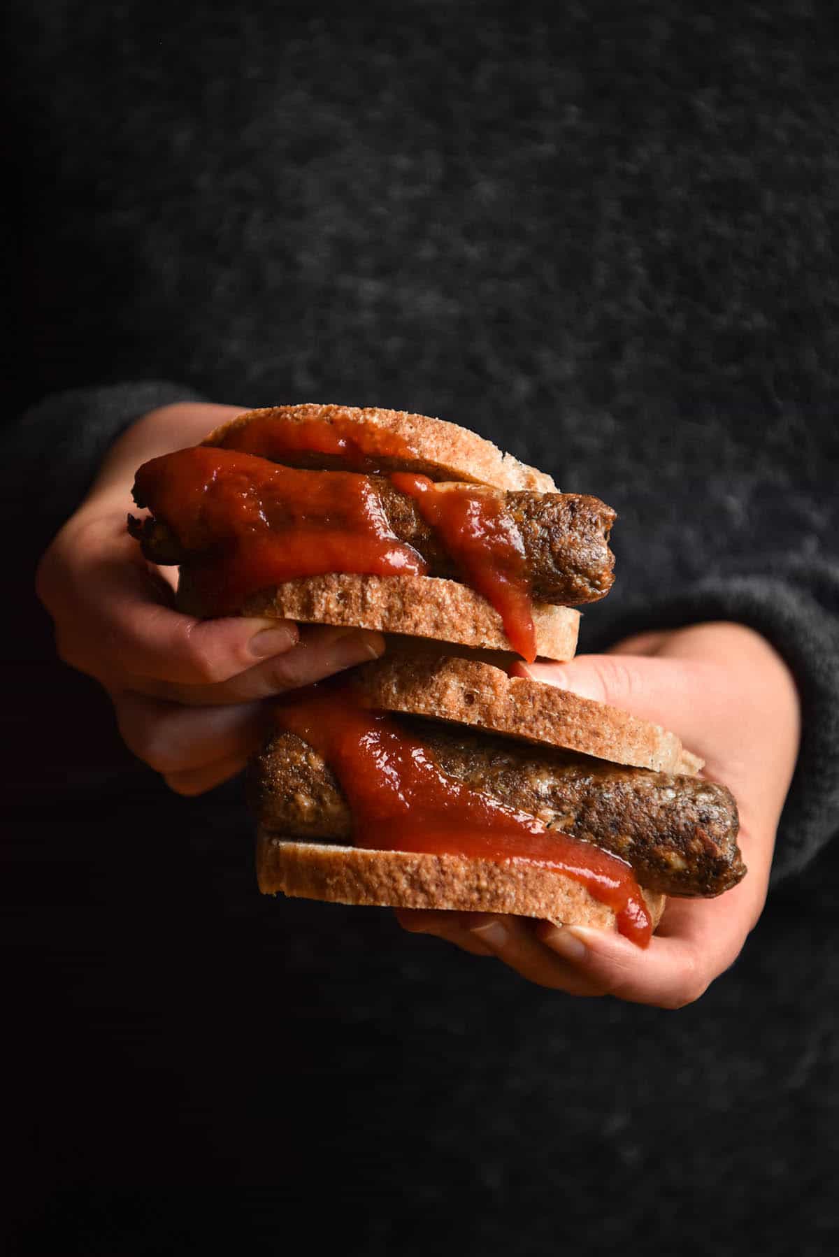 A side on image of a person in a wooly grey jumper holding out two sausage sandwiches made with grain free bread and vegan sausages. The sausages are smothered with tomato sauce.