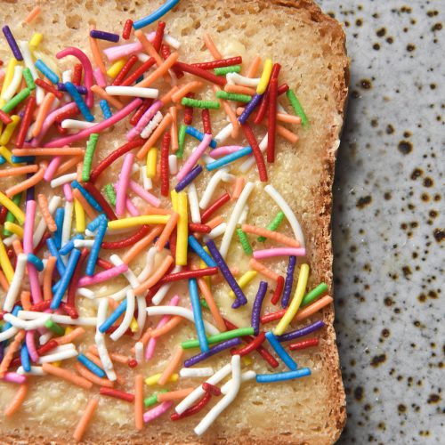 An aerial close up image of a slice of fairybread (white bread topped with butter and sprinkles) on grain free bread. The bread sits atop a white ceramic speckled plate.