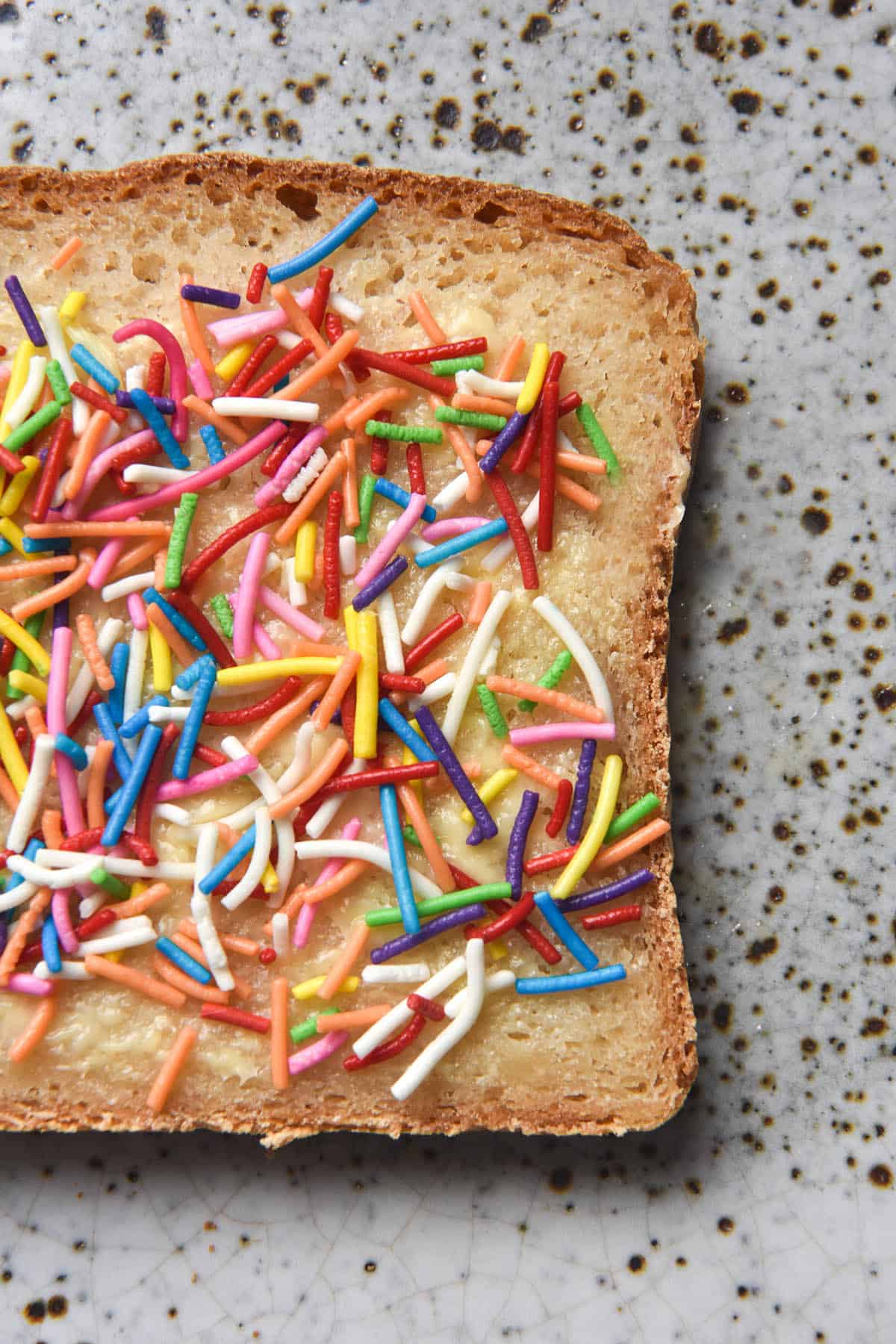 An aerial close up image of a slice of fairybread (white bread topped with butter and sprinkles) on grain free bread. The bread sits atop a white ceramic speckled plate.