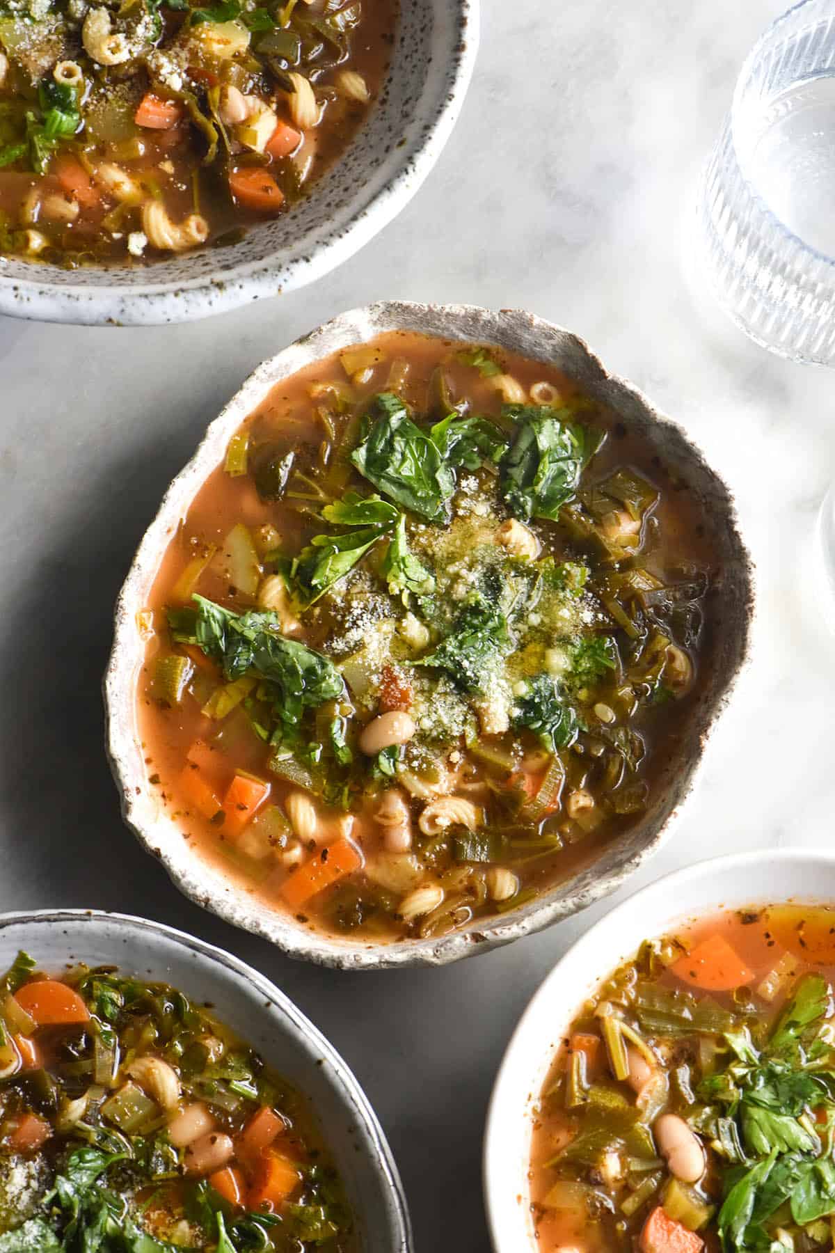 An aerial image of four white ceramic bowls filled with low FODMAP minestrone soup on a white marble table. The bowls are topped with fresh herbs, parmesan and garlic infused olive oil. 