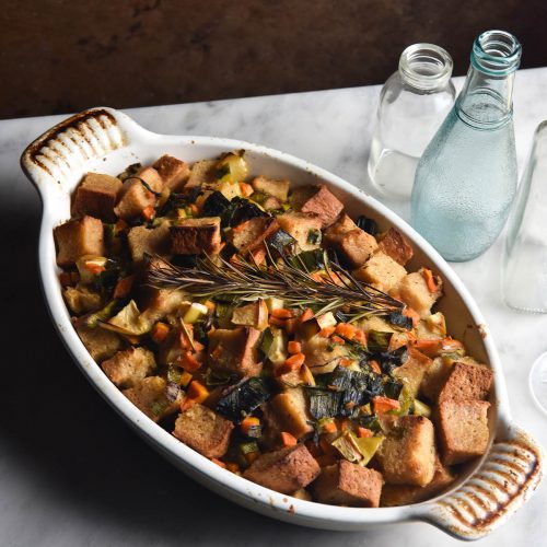 A baking dish filled with low FODMAP stuffing sits on a white marble table against a rusty auburn backdrop. The stuffing is golden brown and topped with a sprig of rosemary. Various coloured water glasses sit to the right of the baking dish.