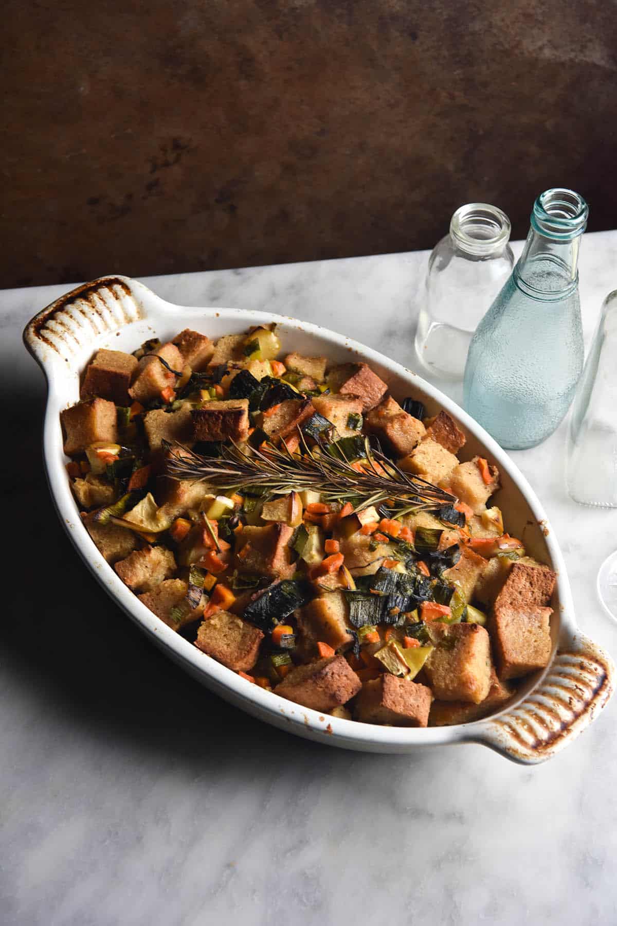 A baking dish filled with low FODMAP stuffing sits on a white marble table with a rusty auburn backdrop. The stuffing is topped with a sprig of rosemary and various coloured glasses sit to the right of the baking dish. 