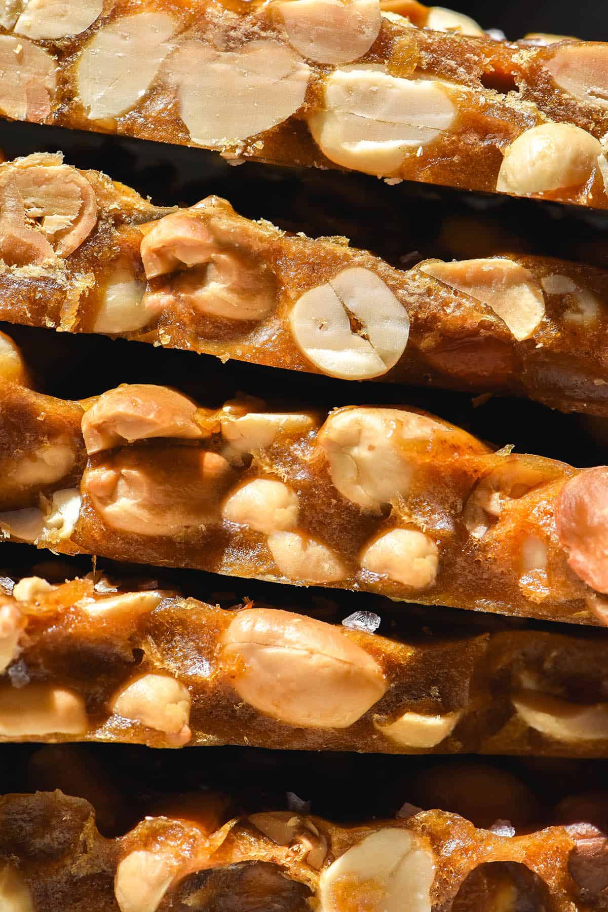 A close up macro image of slices of peanut brittle without corn syrup stacked against a black backdrop. The brittle is filled with roasted peanuts, and slices are topped with flaky sea salt.  