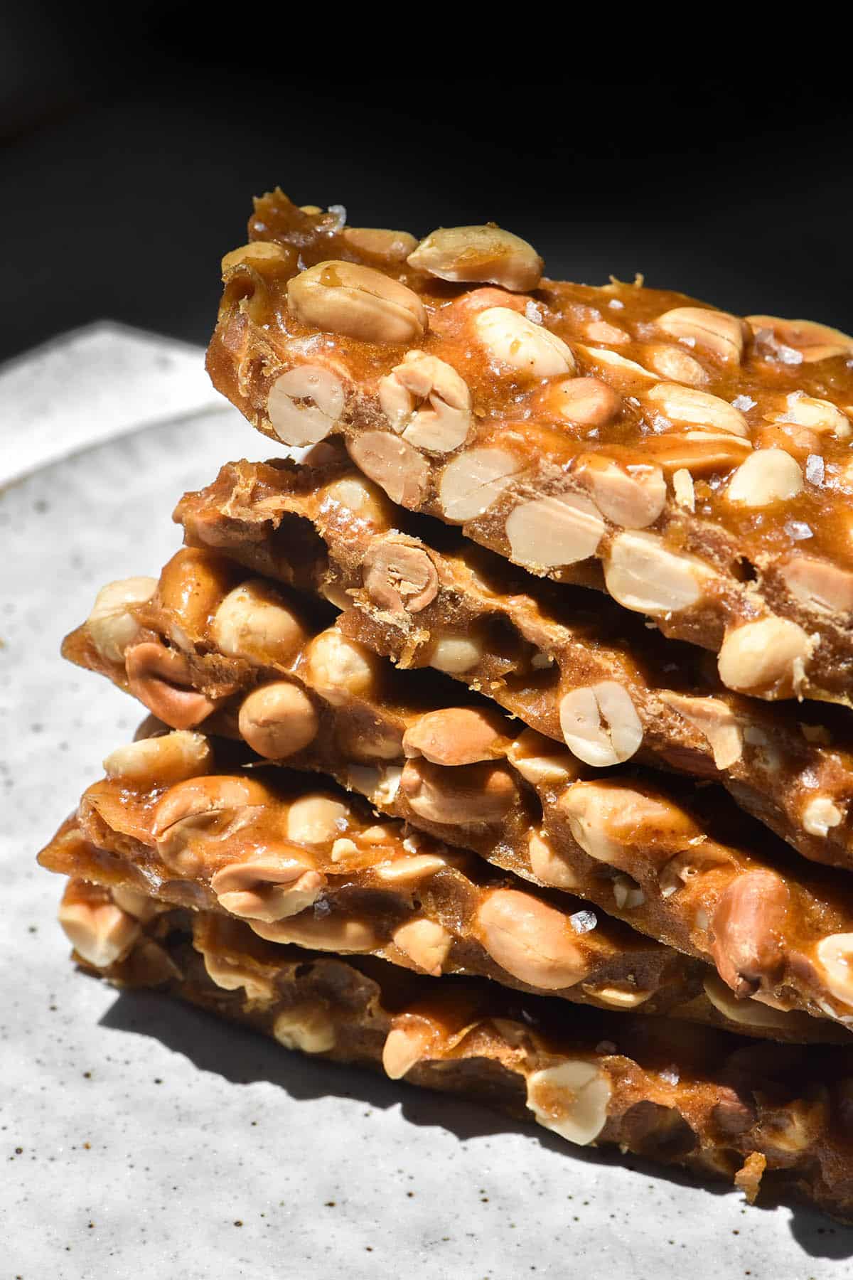 A side on view of a stack of pieces of peanut brittle without corn syrup. The brittle is filled with roasted peanuts and topped with flaky sea salt. It sits atop a white speckled ceramic plate in harsh sunlight, creating dark shadows at the top of the image