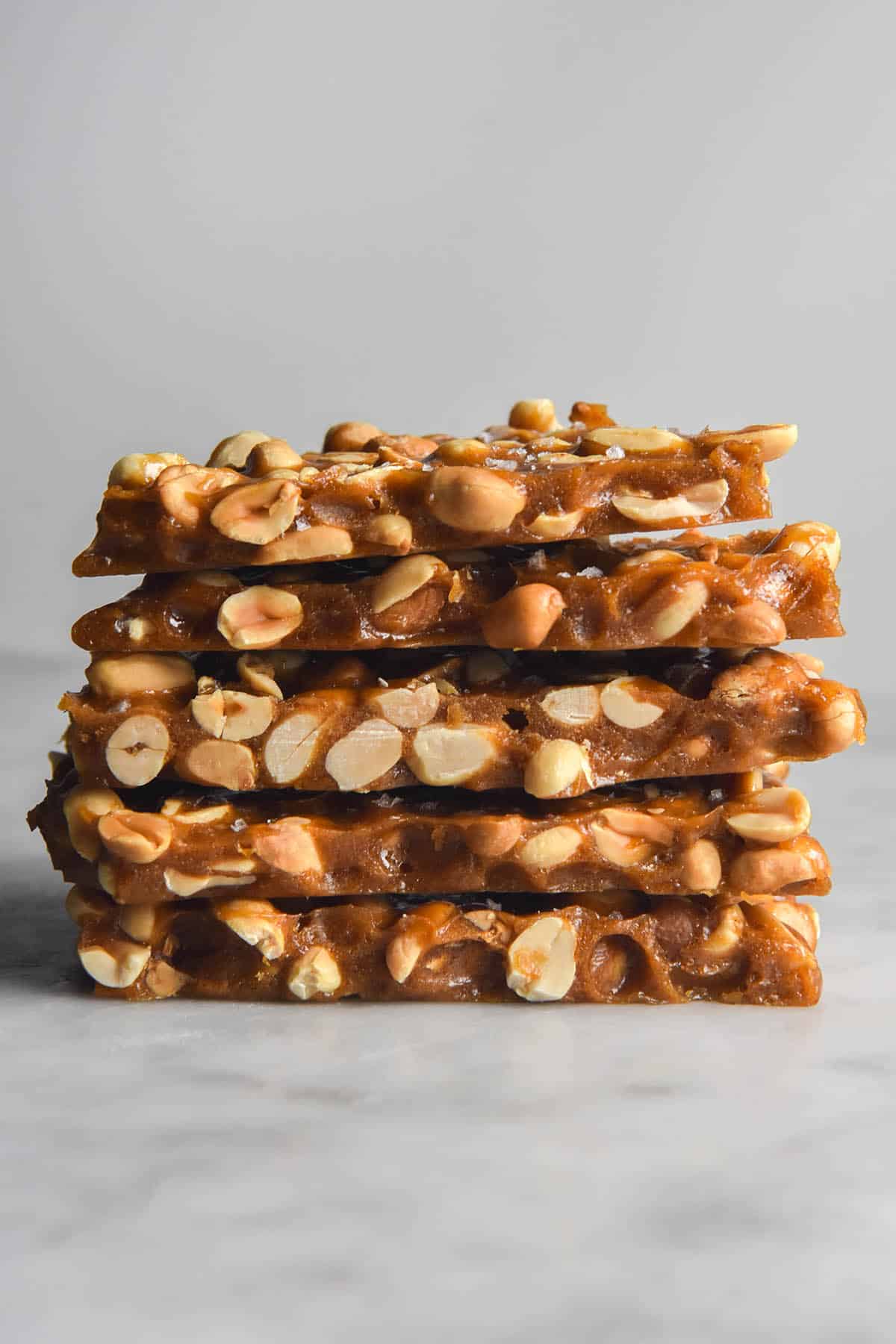 A side on view of 5 slabs of peanut brittle stacked on top of each other on a white table against a white backdrop. 