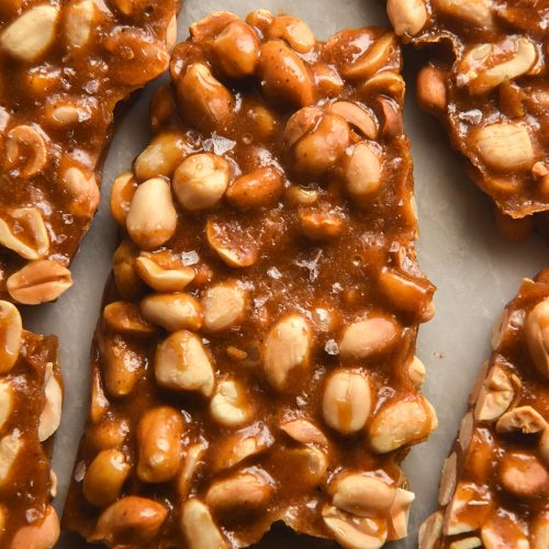 A close up aerial image of slabs of snapped peanut brittle without corn syrup arranged on a white marble table. The centre slab is sprinkled with flaky sea salt