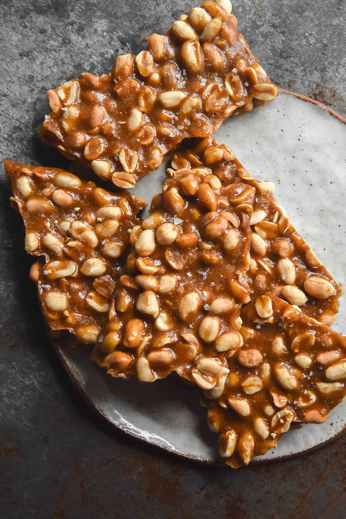 An aerial view of variously sized slabs of peanut brittle without corn syrup on a small white ceramic plate on a dark steel backdrop. The brittle is golden brown, casually arranged and sprinkled with flaky sea salt