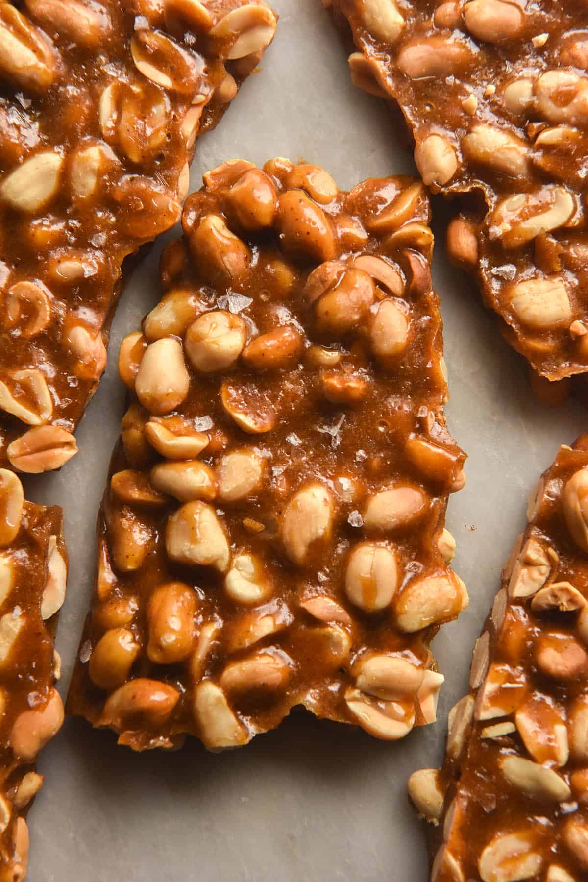 A close up aerial image of slabs of snapped peanut brittle without corn syrup arranged on a white marble table. The centre slab is sprinkled with flaky sea salt