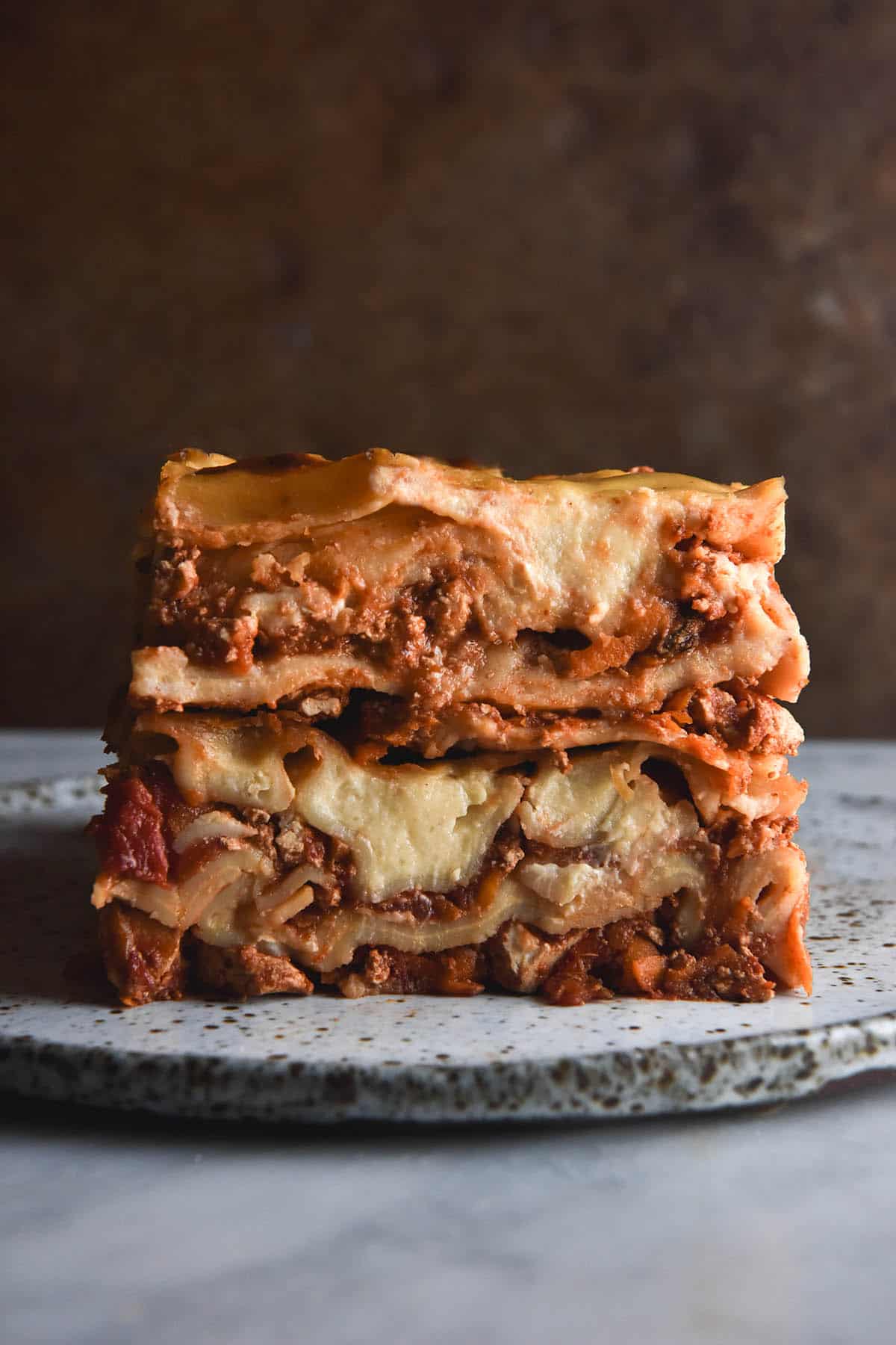 A side on image of a stack of gluten free vegan lasagne on a white speckled plate against a dark rust coloured backdrop