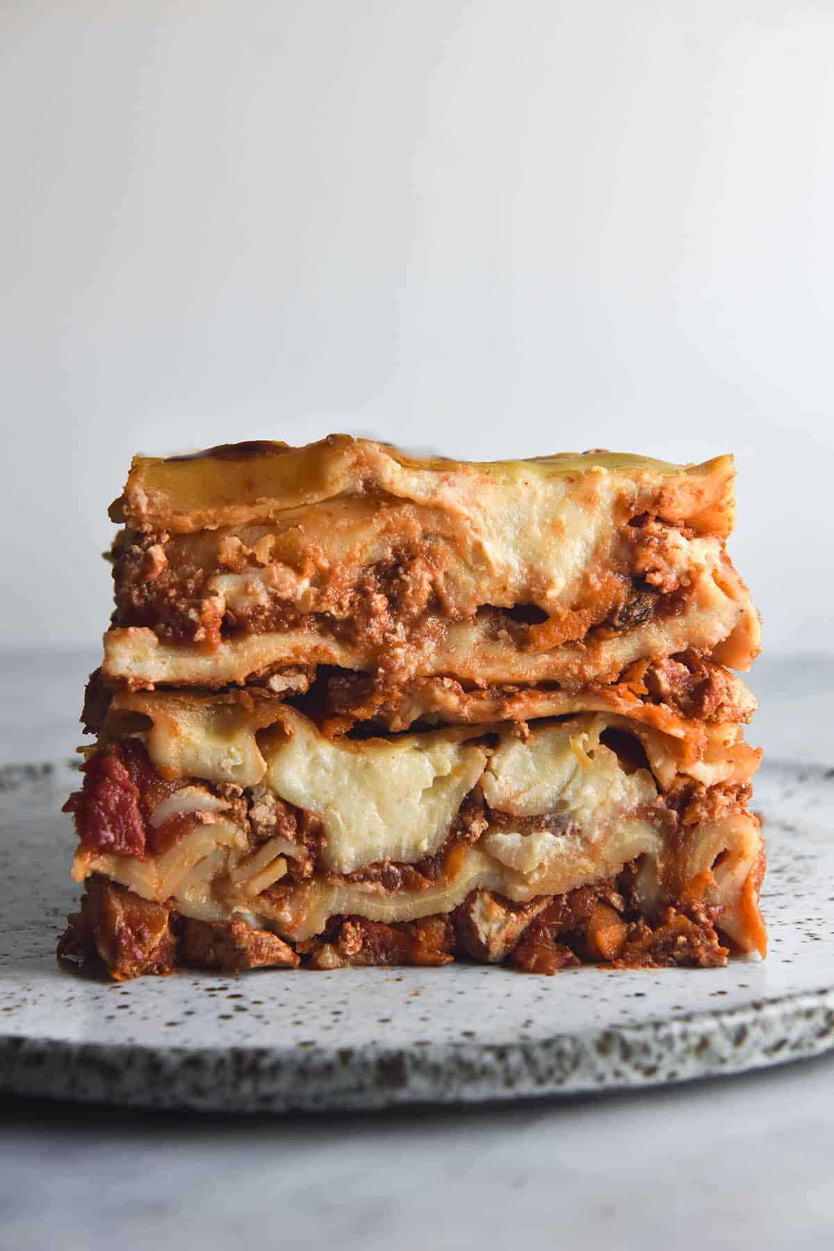 A side on view of a stack of gluten free vegan lasagne. The stack sits on a white speckled ceramic plate on a white marble table. It sits against a white backdrop. 