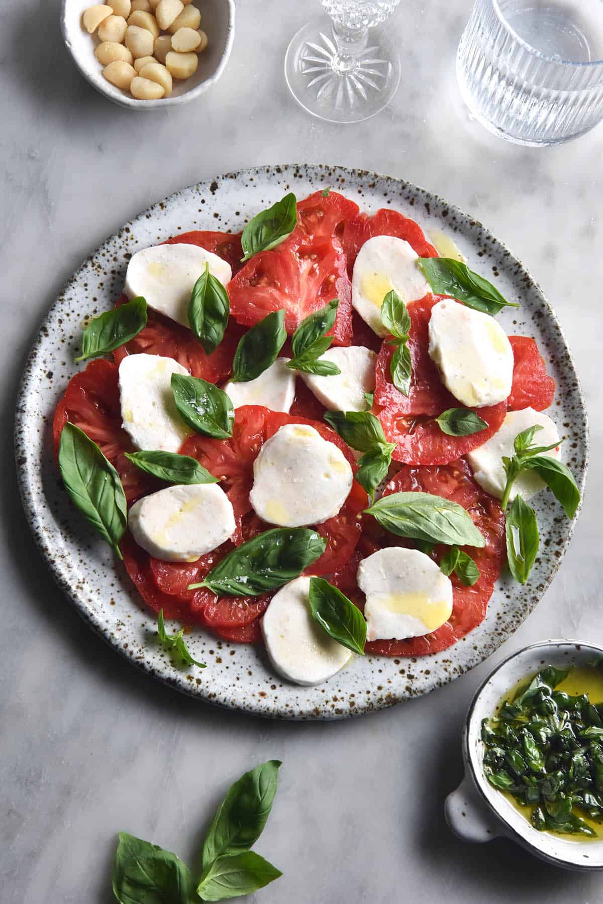 An aerial image of a caprese salad on a white marble table. The caprese is surrounded by extra tomatoes, basil and water glasses. The mozzarella used in the salad is vegan mozzarella made from macadamias.