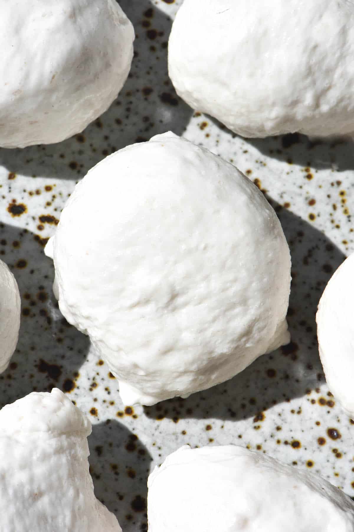 Balls of homemade vegan mozzarella sit on a white speckled ceramic plate in harsh sunlight. The image is a close up aerial image and shows the creamy white texture of the mozzarella.