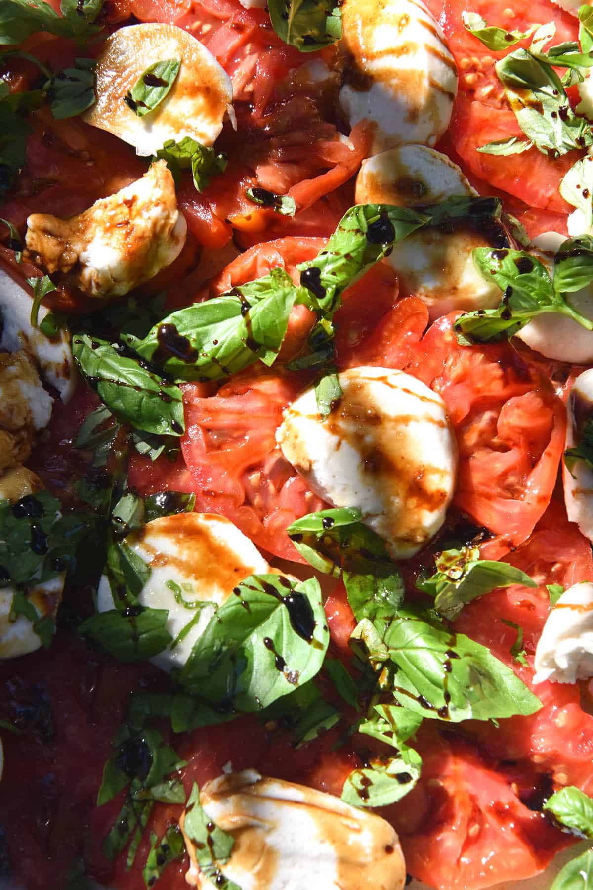 An aerial sunlit view of a caprese salad with vegan mozzarella. The caprese is finished with a balsamic reduction drizzled over, which creates a dramatic pattern and a golden orange hue on the mozzrella