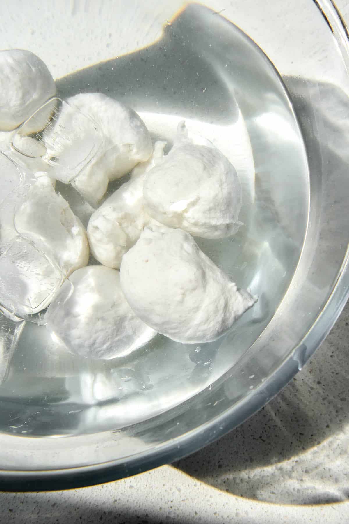 A close up aerial image of balls of vegan mozzarella in a bath of ice water. The bowl sits in the sunlight on a white speckled stone bench