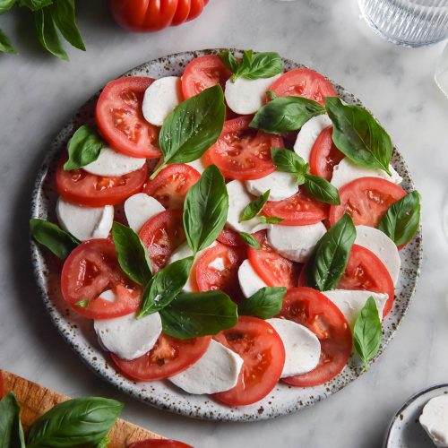 An aerial image of a caprese salad on a white marble table. The caprese is surrounded by extra tomatoes, basil and water glasses. The mozzarella used in the salad is vegan mozzarella made from macadamias.