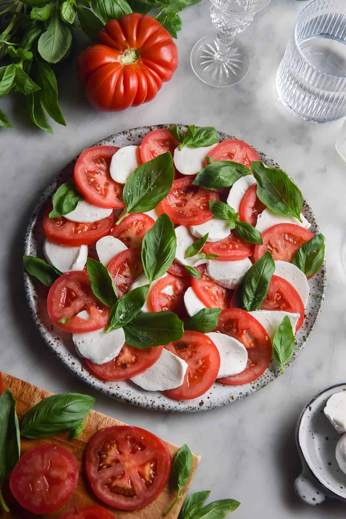 An aerial image of a caprese salad on a white marble table. The caprese is surrounded by extra tomatoes, basil and water glasses. The mozzarella used in the salad is vegan mozzarella made from macadamias.