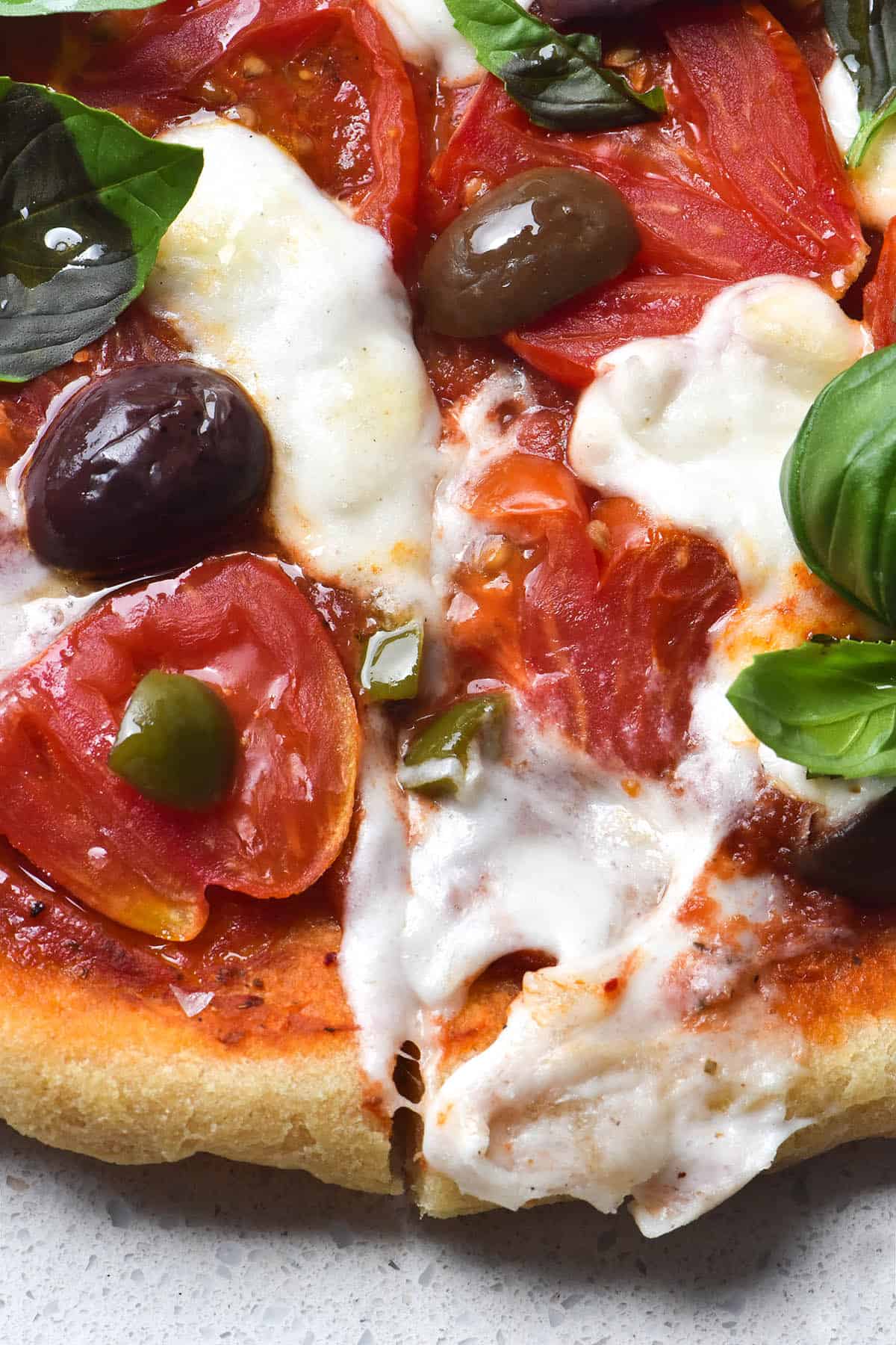 A close up view of a pizza topped with pizza sauce, olives, heirloom tomatoes, basil and melty vegan mozzarella. The pizza has a slice down the centre and sits on a white stone benchtop.