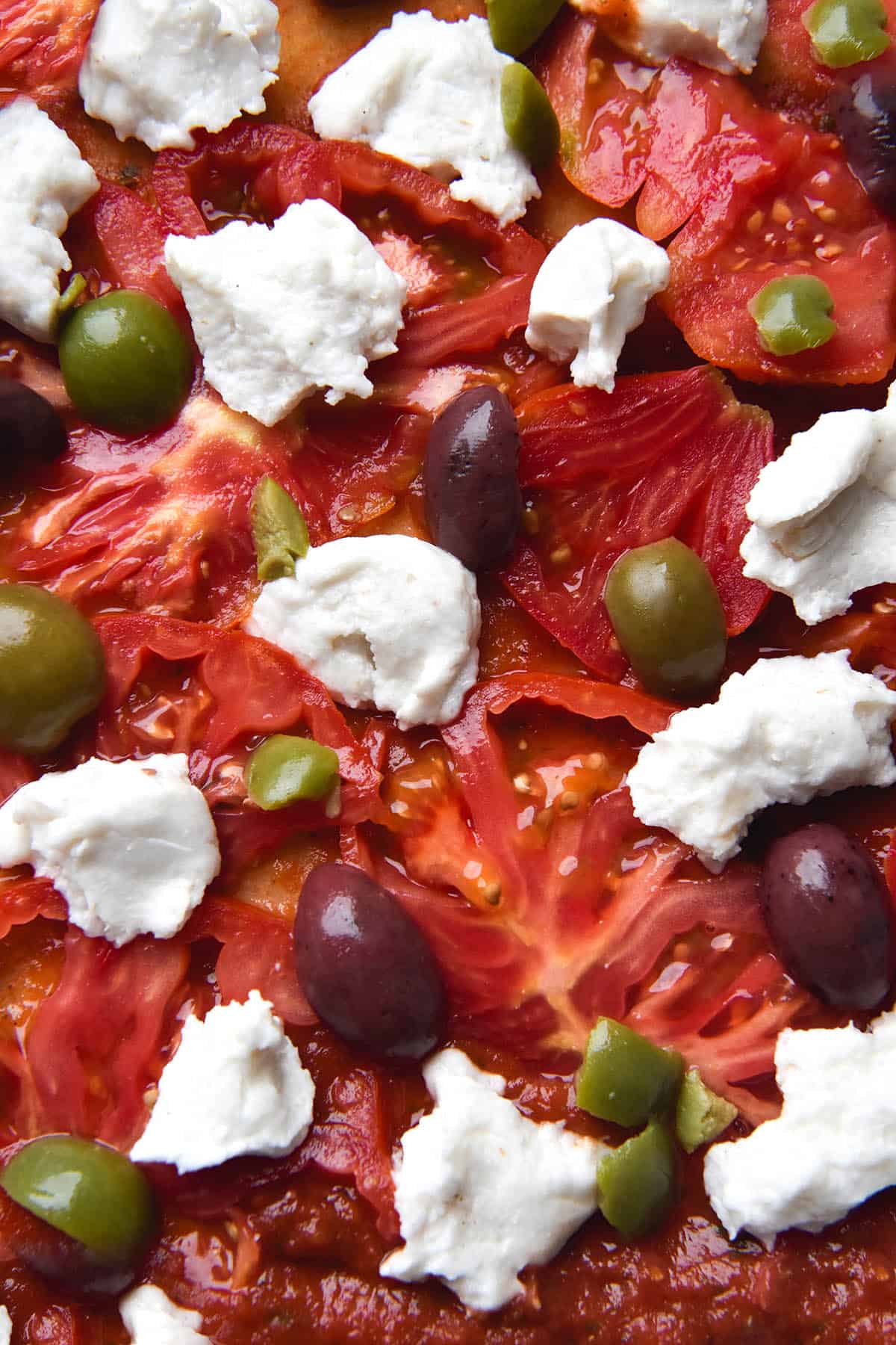 An aerial close up view of a pizza topped with pizza sauce, sliced heirloom tomatoes, different varieties of olives and vegan mozzarella.