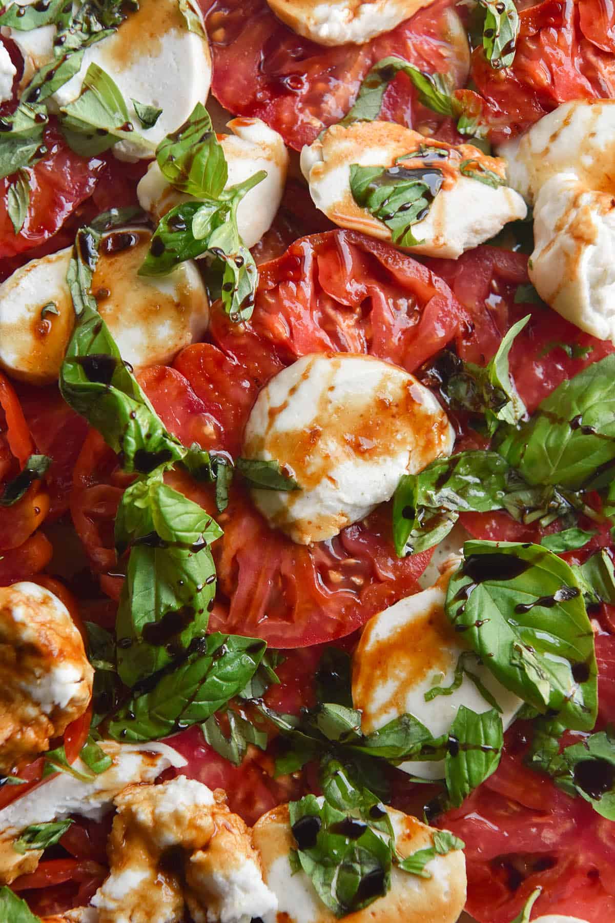 An aerial close up image of vegan macadamia mozzarella in a caprese salad. The salad is drizzled with a balsamic reduction which colours parts of the mozzarella in a deep orange hue