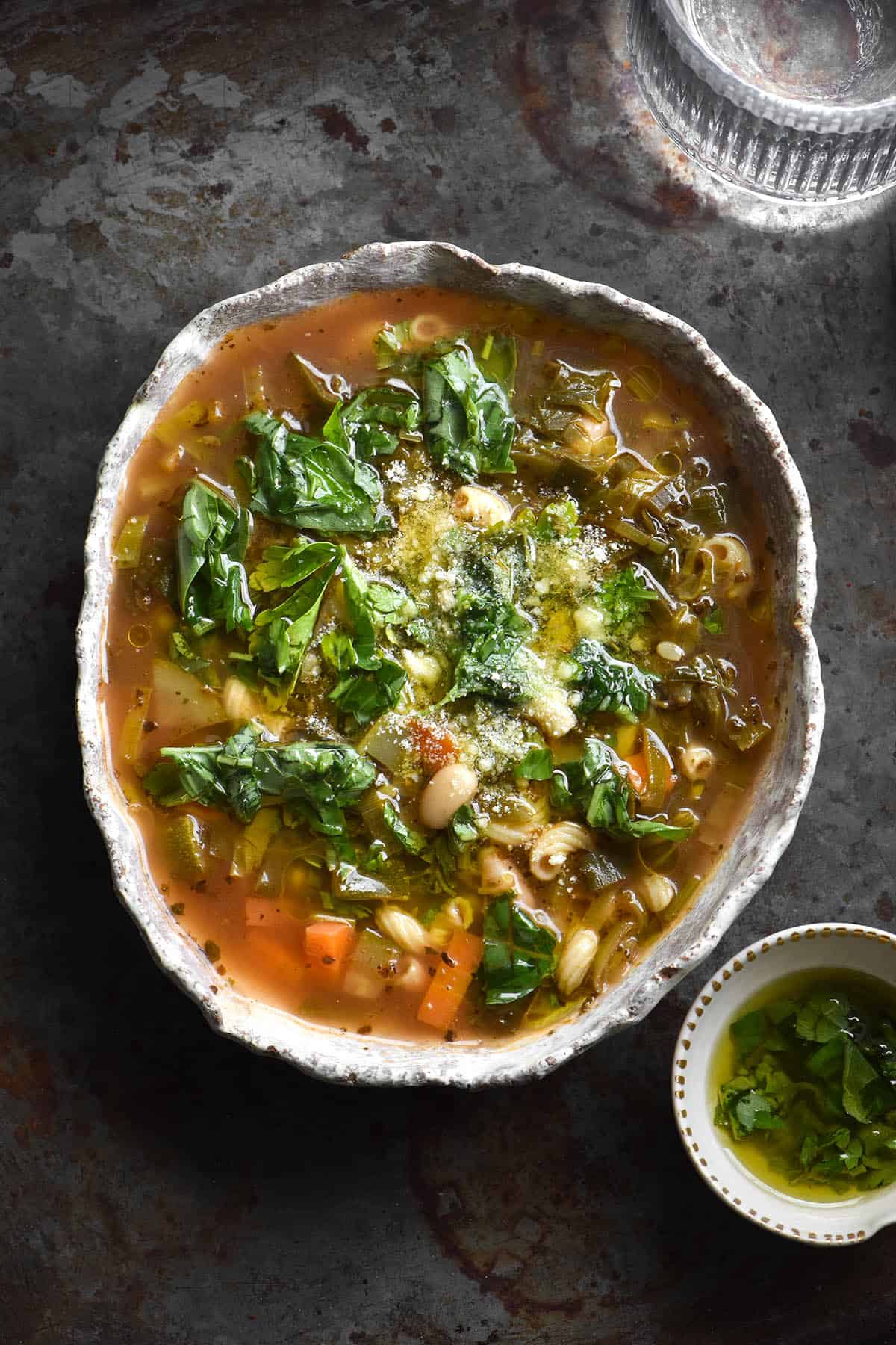 An aerial view of a white misshapen ceramic bowl filled with low FODMAP minestrone soup. The soup is topped with garlic infused oil, herbs and finely grated parmesan. The bowl sits on a dark grey steel backdrop and a small bowl of herb oil sits to the bottom right of the image, while a water glass sits to the top right. 