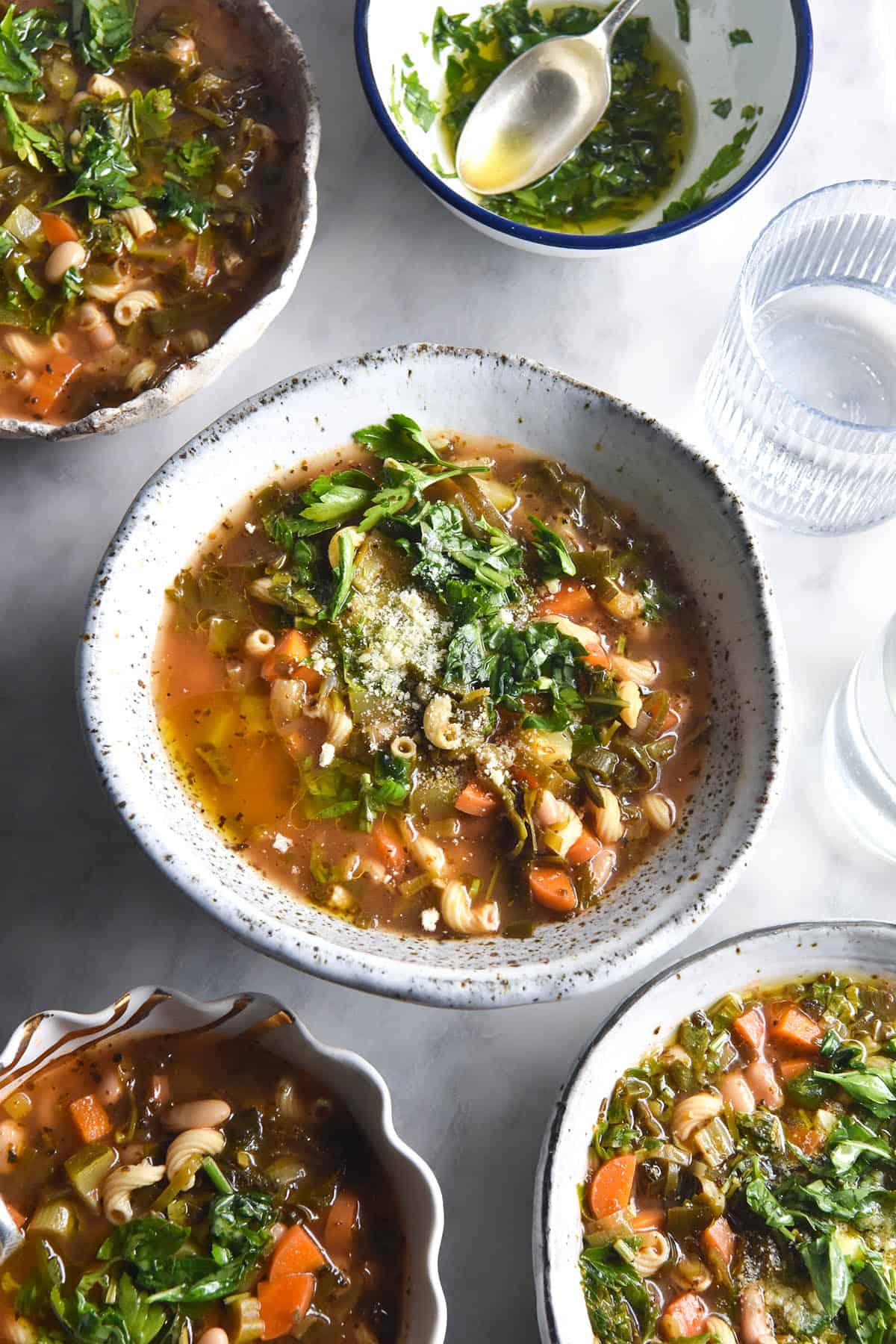 An aerial image of various white ceramic bowls filled with low FODMAP minestrone arranged on a white marble table. The bowls are topped with fresh herbs, parmesan and garlic infused olive oil. The bowls surround a centre bowl, and glasses of water sit to the right hand side of the centre bowl