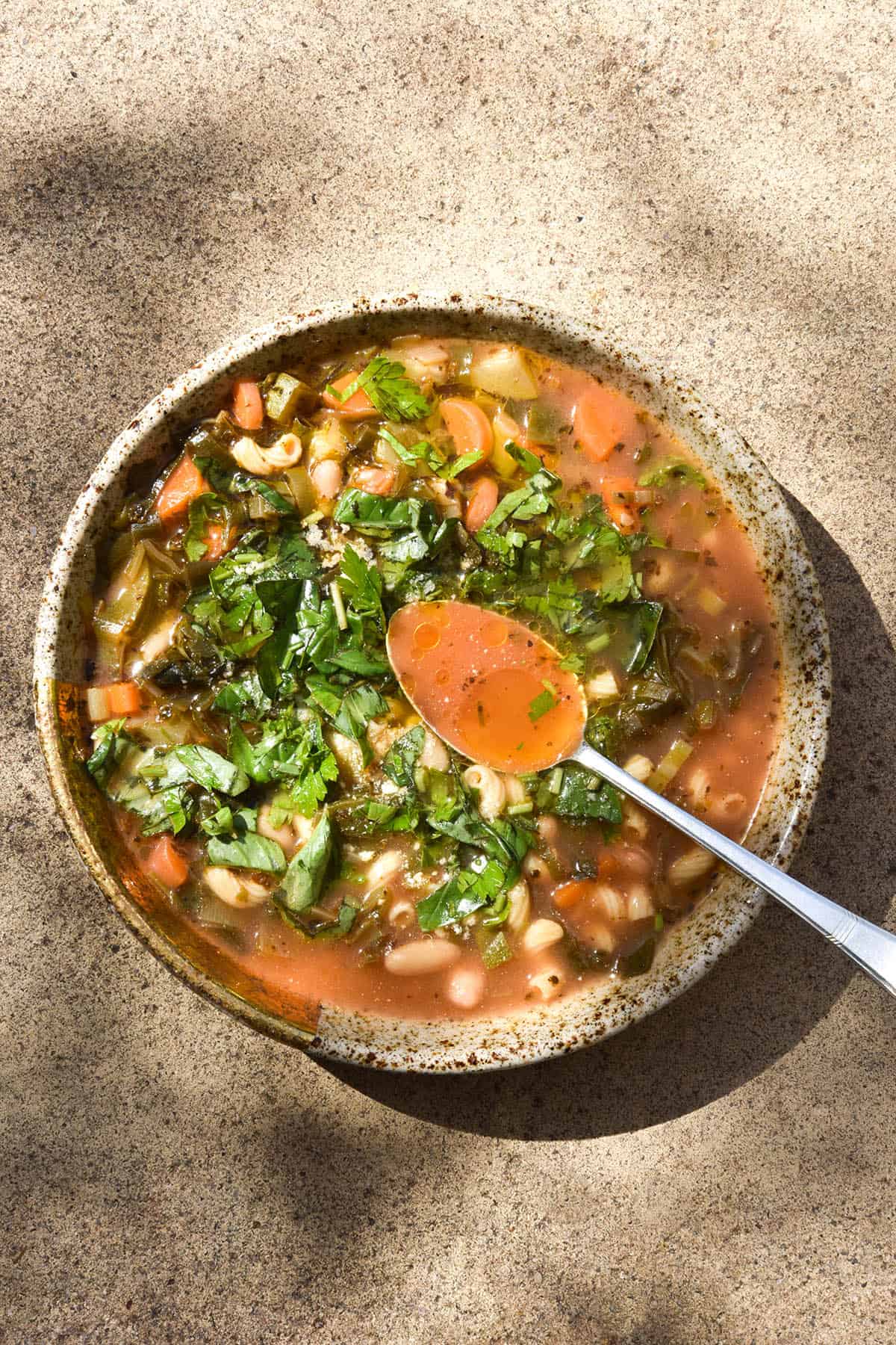 An aerial image of a beige speckled ceramic bowl of low FODMAP minestrone sitting on a stone backdrop in dappled sunlight. The minestrone is topped with herbs and garlic infused olive oil. A spoon dips into the minestrone from the bottom right hand side of the image
