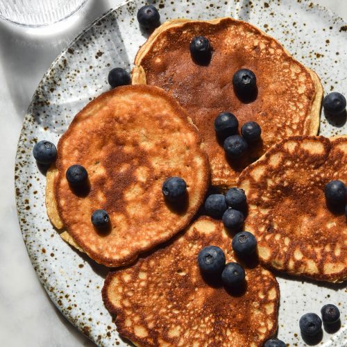 An aerial image of a plate of buckwheat banana pancakes on a white marble table. The pancakes are topped with blueberries and two glasses of water sit in the top of the image.