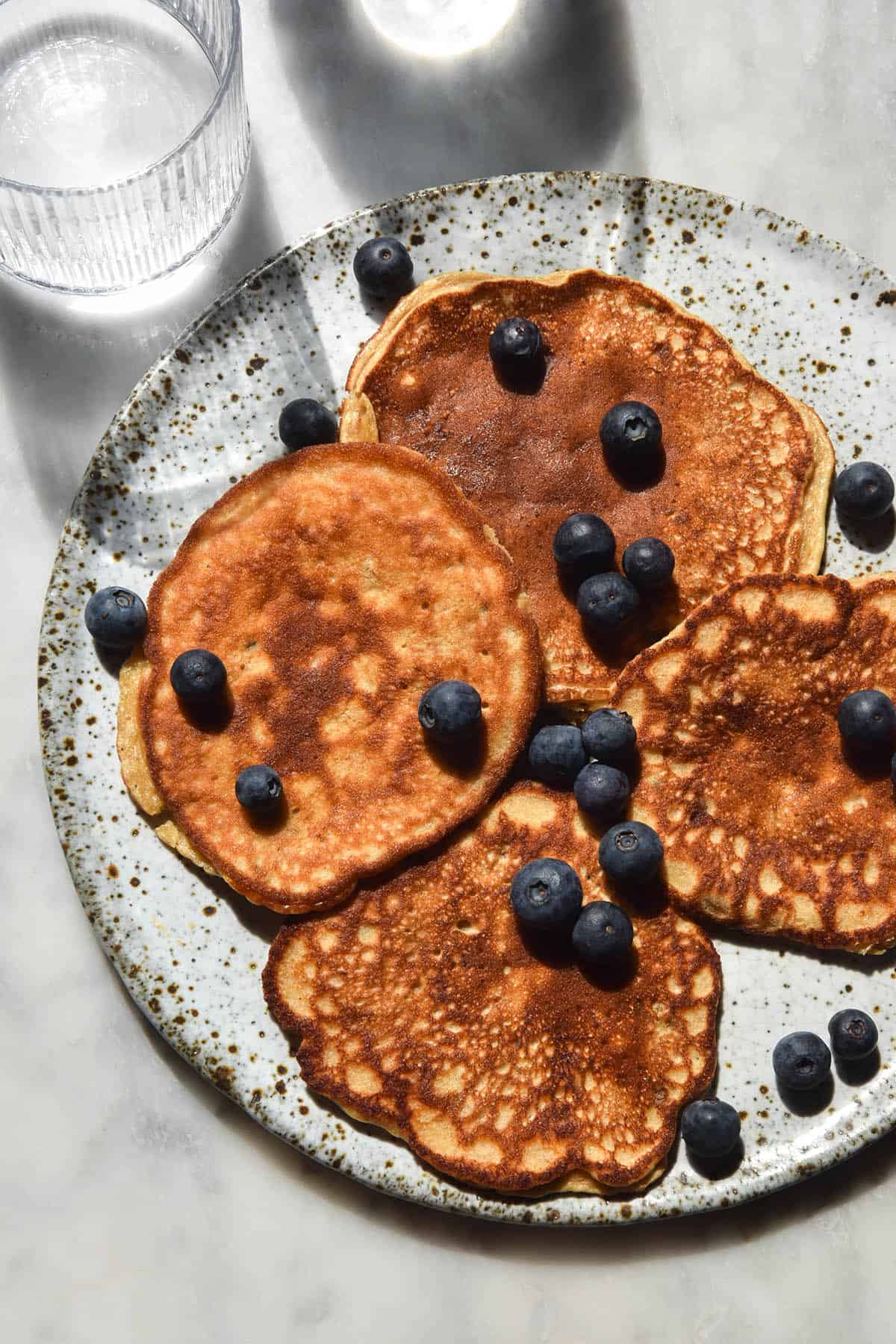 An aerial close up view of banana protein pancakes on a white speckled ceramic plate atop a white marble table. The pancakes are in bright sunlight and water glasses in the top of the image cast light across the plate. The pancakes are scattered with some blueberries. 