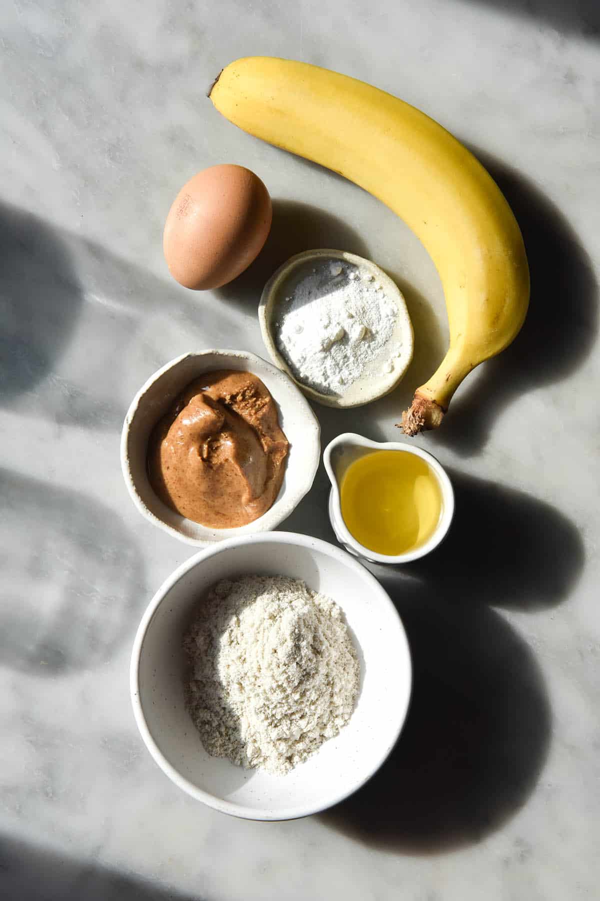 An aerial view of the ingredients for protein powder pancakes in small ceramic pinch bowls on a white marble table in harsh sunlight. Water glasses sit to the left of the image and cast shadow patterns across the image. 