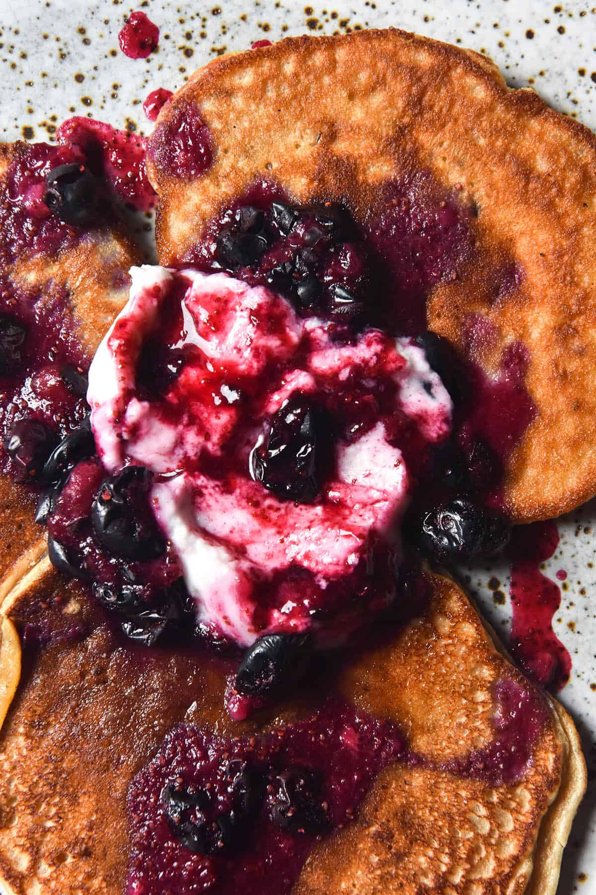 An aerial close up view of banana protein pancakes smothered in coconut yoghurt and blueberry compote. The compote mixes with the yoghurt to create a pink and purple marbled effect