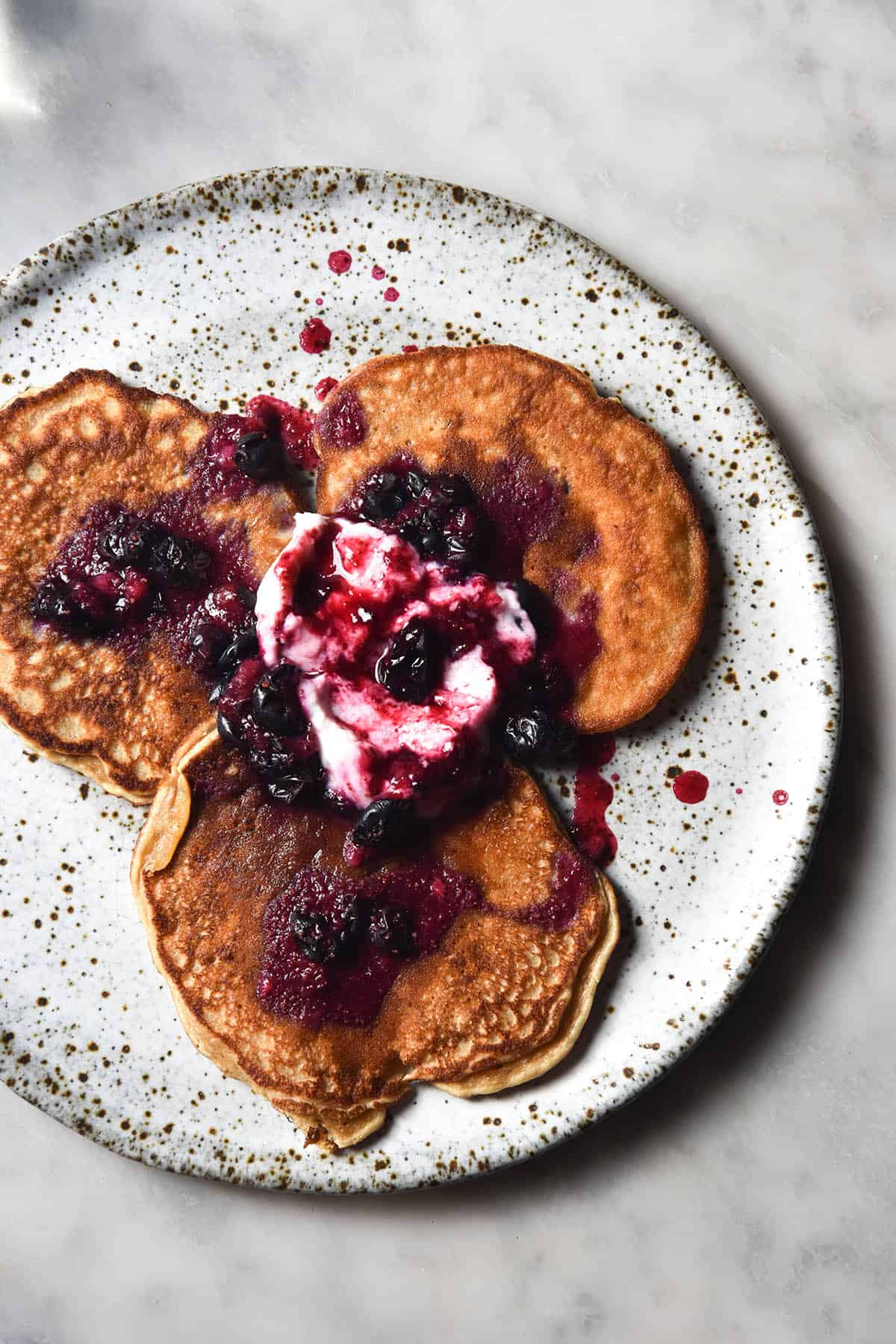 An aerial view of a plate of banana protein pancakes on a white speckled ceramic plate against a white marble table. The pancakes are smothered in coconut yoghurt and blueberry compote which mingle together to create a bright pink colour