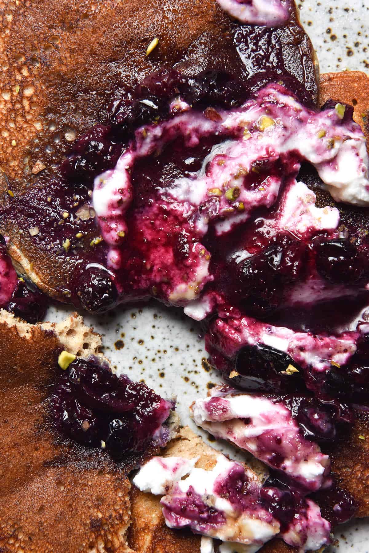 An aerial close up view of some sliced up banana protein pancakes topped with a messy combination of blueberry compote and coconut yoghurt. The pancake has been cut across the middle on an angle, revealing the ceramic speckled plate underneath. 