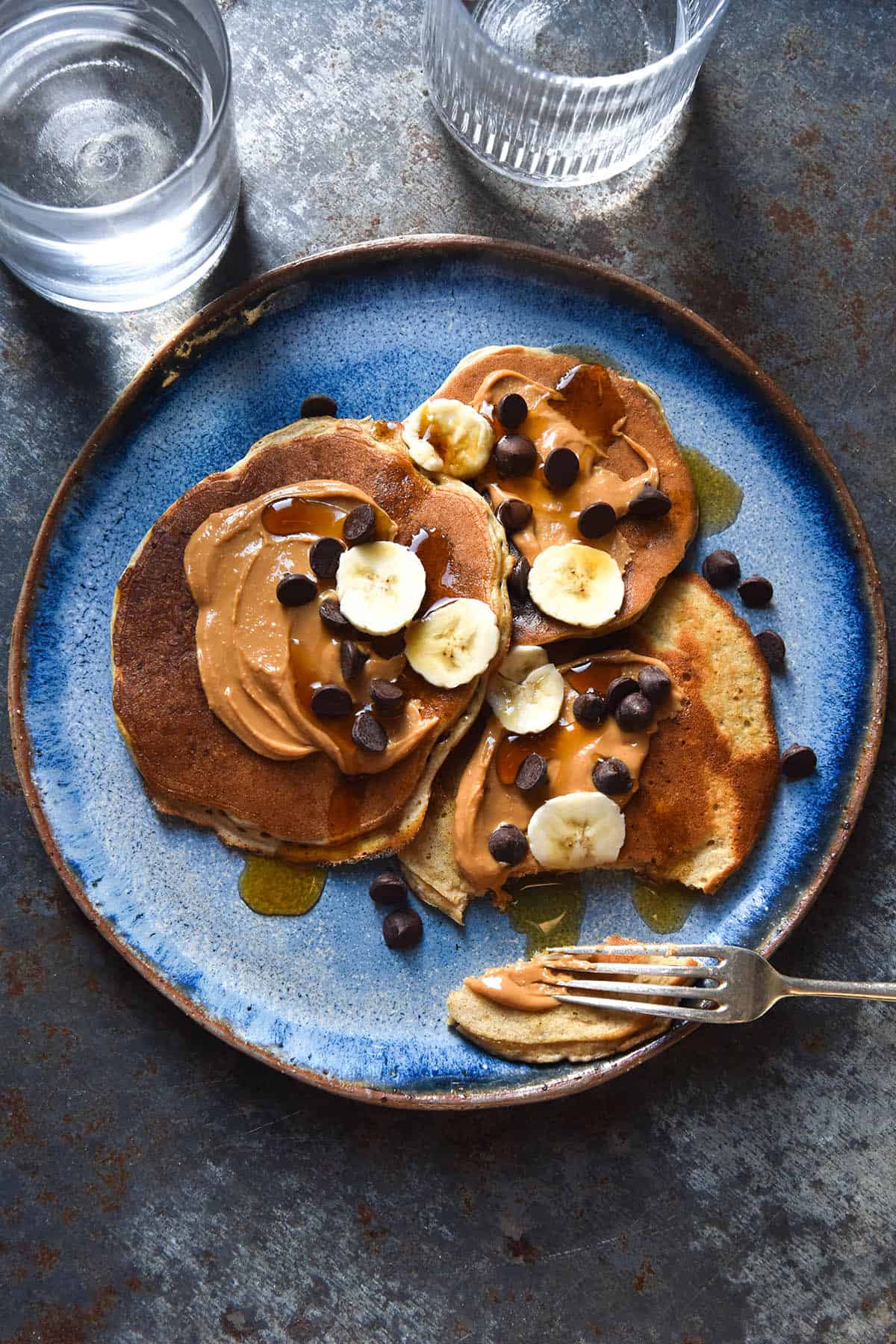 An aerial view of a bright blue ceramic plate topped with banana protein pancakes smothered in peanut butter and finished with choc chips, banana coins and maple syrup. The plate sits atop a mid blue steel backdrop.