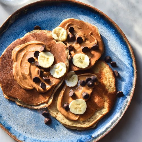 An aerial view of a bright blue ceramic plate topped with gluten free protein pancakes smothered in peanut butter and topped with banana coins, choc chips and maple syrup. The plate sits on a white marble table that is in filtered sunlight.