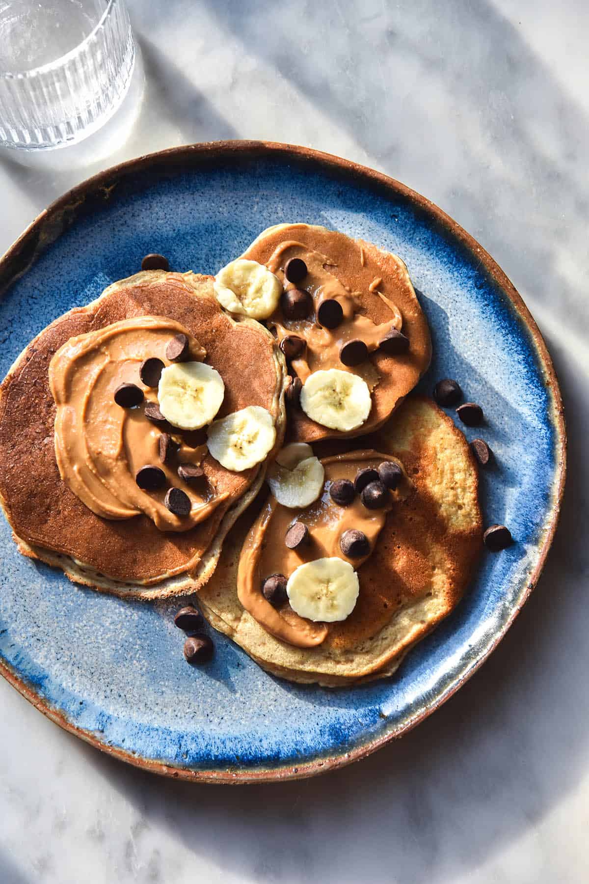 An aerial view of a bright blue ceramic plate topped with banana protein pancakes smothered in peanut butter and topped with chocolate chips and banana coins. The plate sits on a white marble table in cool sunlight