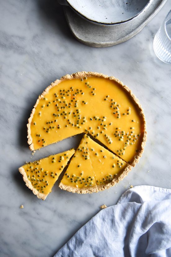 An aerial image of a dairy free lemon curd tart topped with passionfruit seeds on a white marble table. The tart has been sliced and one slice sits at a small distance from the tart.