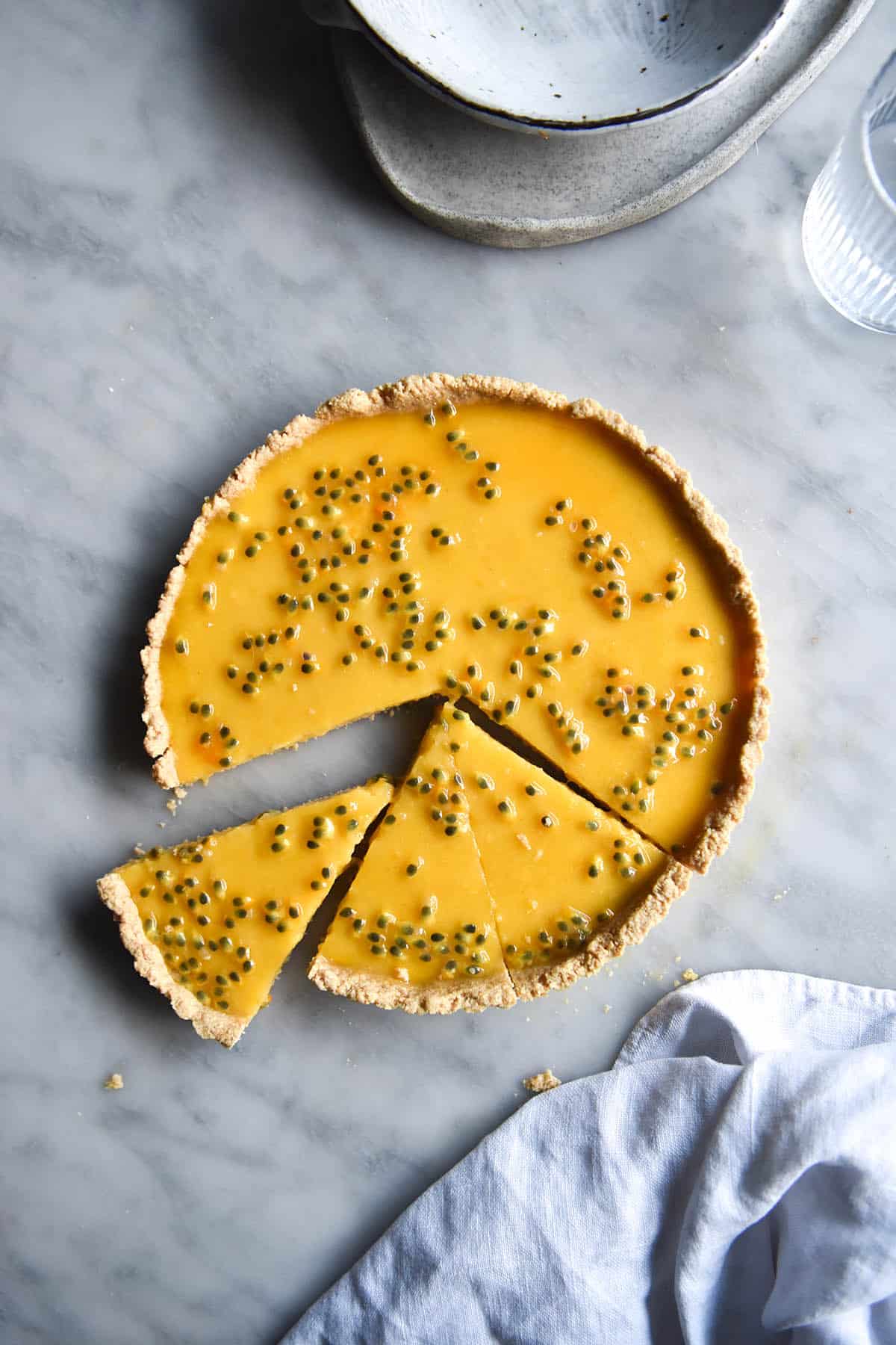 An aerial image of a dairy free lemon curd tart topped with passionfruit seeds on a white marble table. The tart has been sliced and one slice sits at a small distance from the tart. 