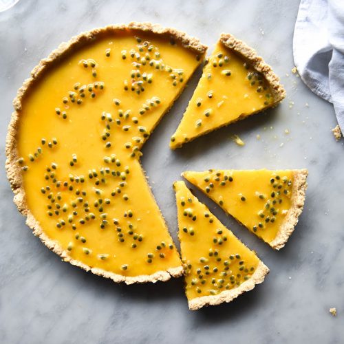 An aerial view of a dairy free lemon curd tart topped with passionfruit on a white marble table. The tart has been sliced and the pieces sit slightly away from the tart. A white linen cloth sits to the right of the tart.