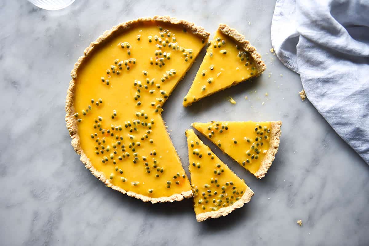 An aerial view of a dairy free lemon curd tart topped with passionfruit on a white marble table. The tart has been sliced and the pieces sit slightly away from the tart. A white linen cloth sits to the right of the tart.