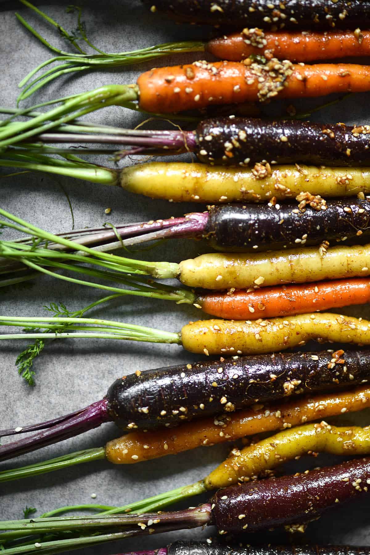 An aerial view of heirloom carrots lined up on a baking tray, smothered in oil and dukkah. The carrots are on the right side of the image and their tops extend off to the left of the image