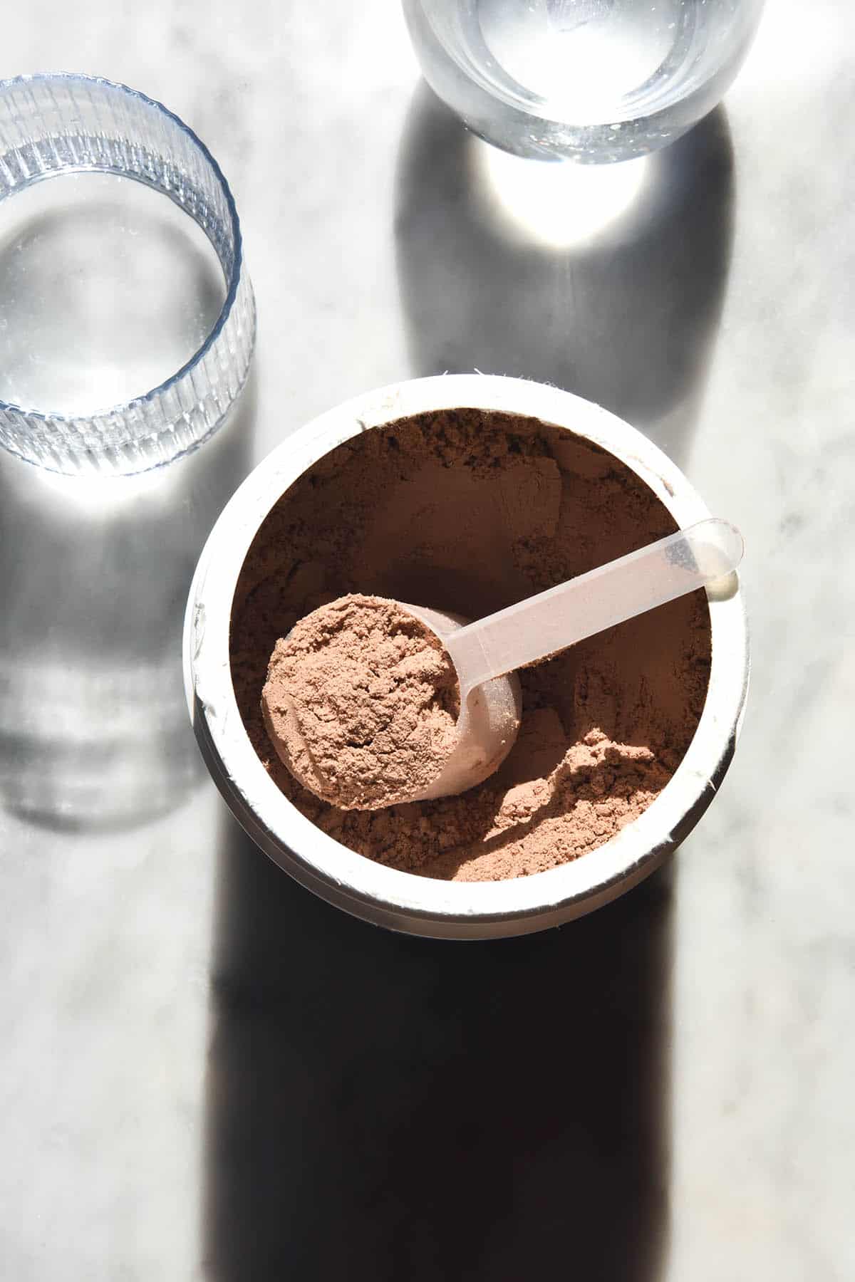 An aerial view of a container of chocolate protein powder with the scoop being removed. The container sits on a white marble table in sunlight with two glasses of water in the background 