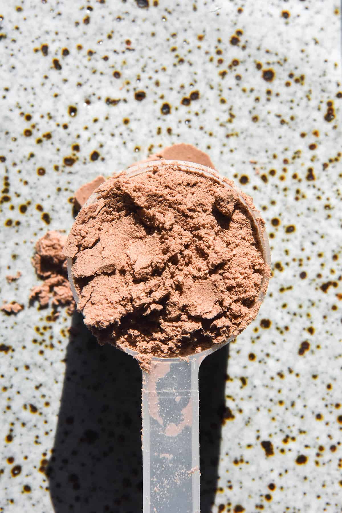 A close up aerial image of a scoop of  chocolate protein powder on a white speckled ceramic plate