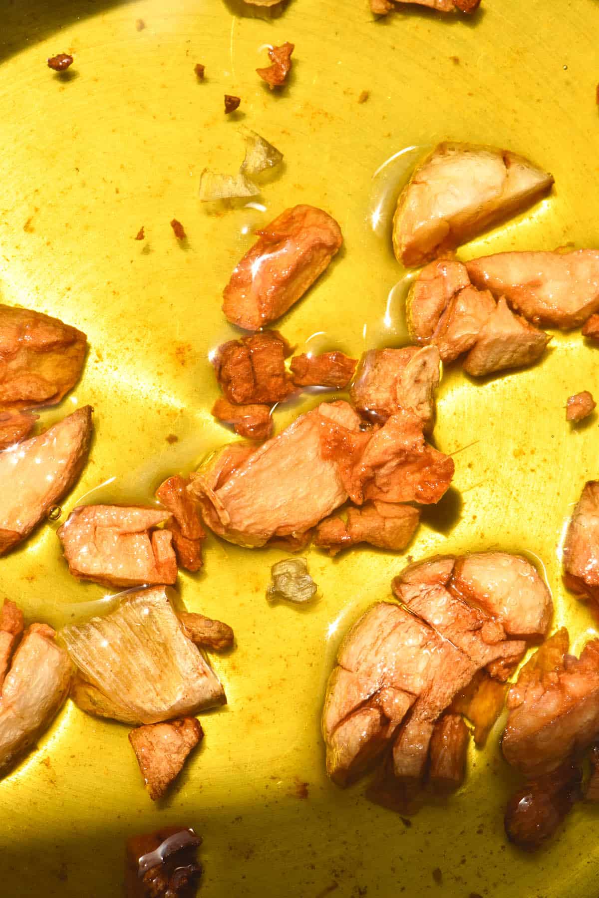 An aerial close up macro shot of a saucepan filled with garlic infused oil in bright sunlight. Pieces of caramelised garlic sit in the bright yellow oil.