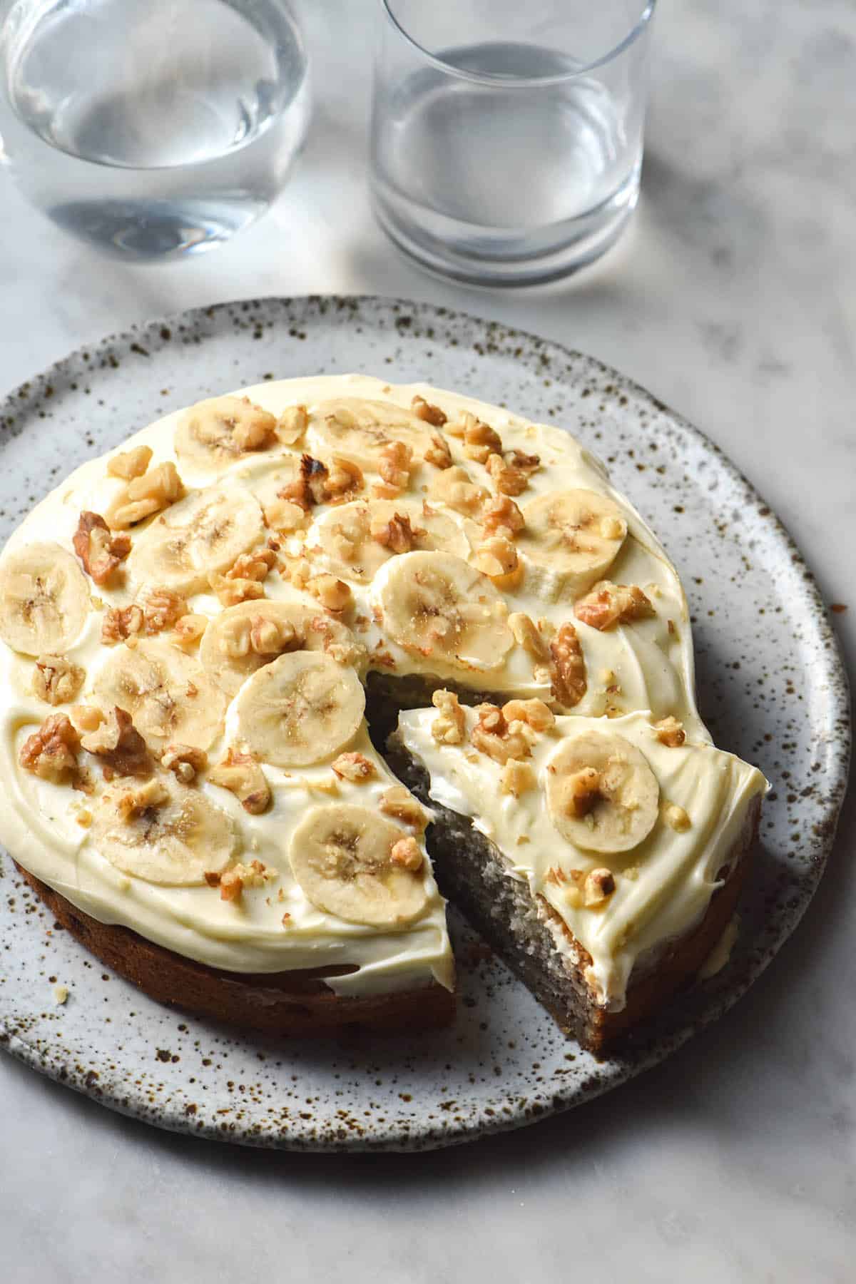 A side on view of a gluten free banana cake on a white speckled ceramic plate. The banana cake is topped with cream cheese icing, sliced banana and crushed walnuts. 