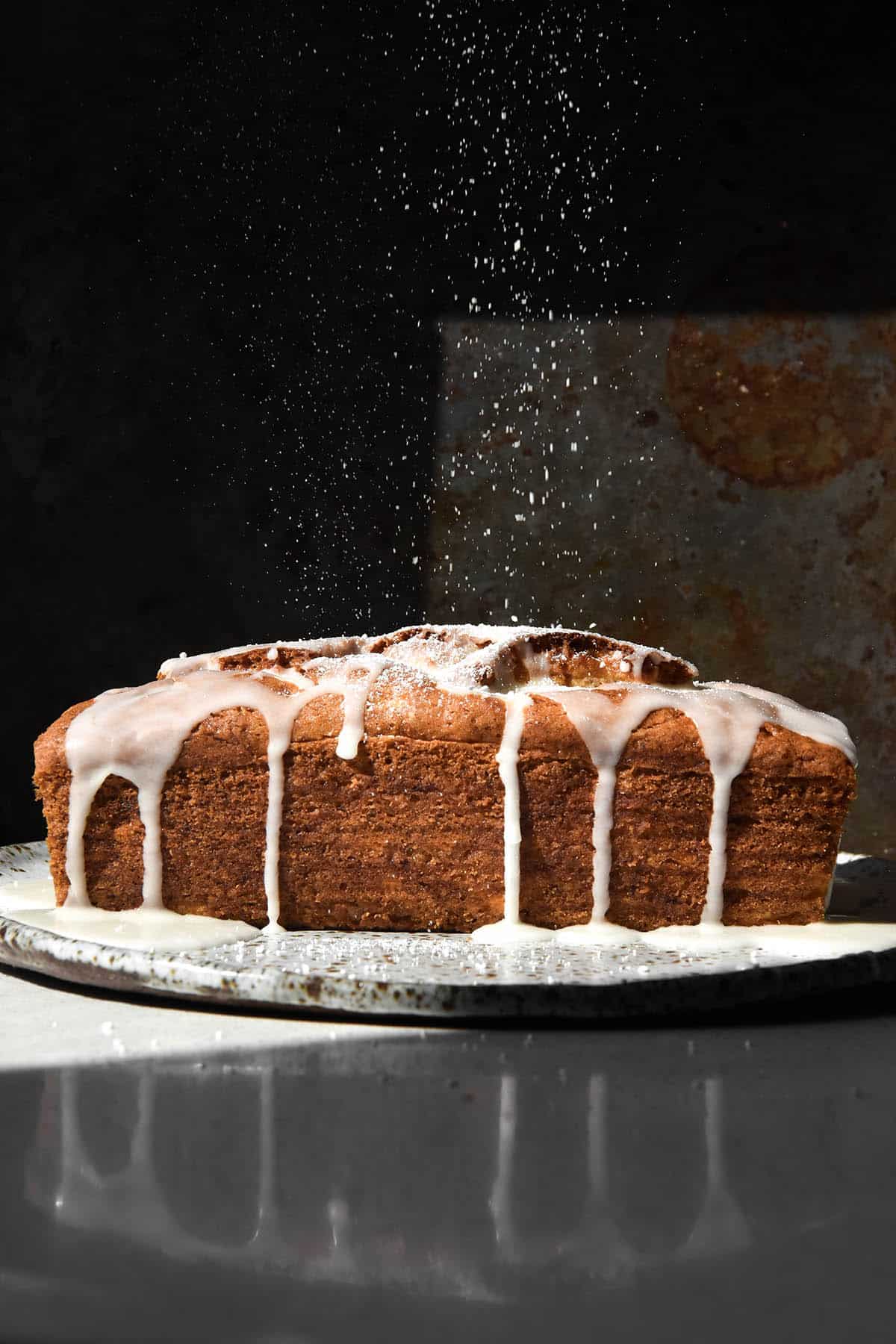 A side on view of a gluten free banana loaf in harsh contrasting light. The loaf is drizzled with a lemon glaze, and a sprinkle of icing sugar drifts down from the top of the image, contrasting against the dark background