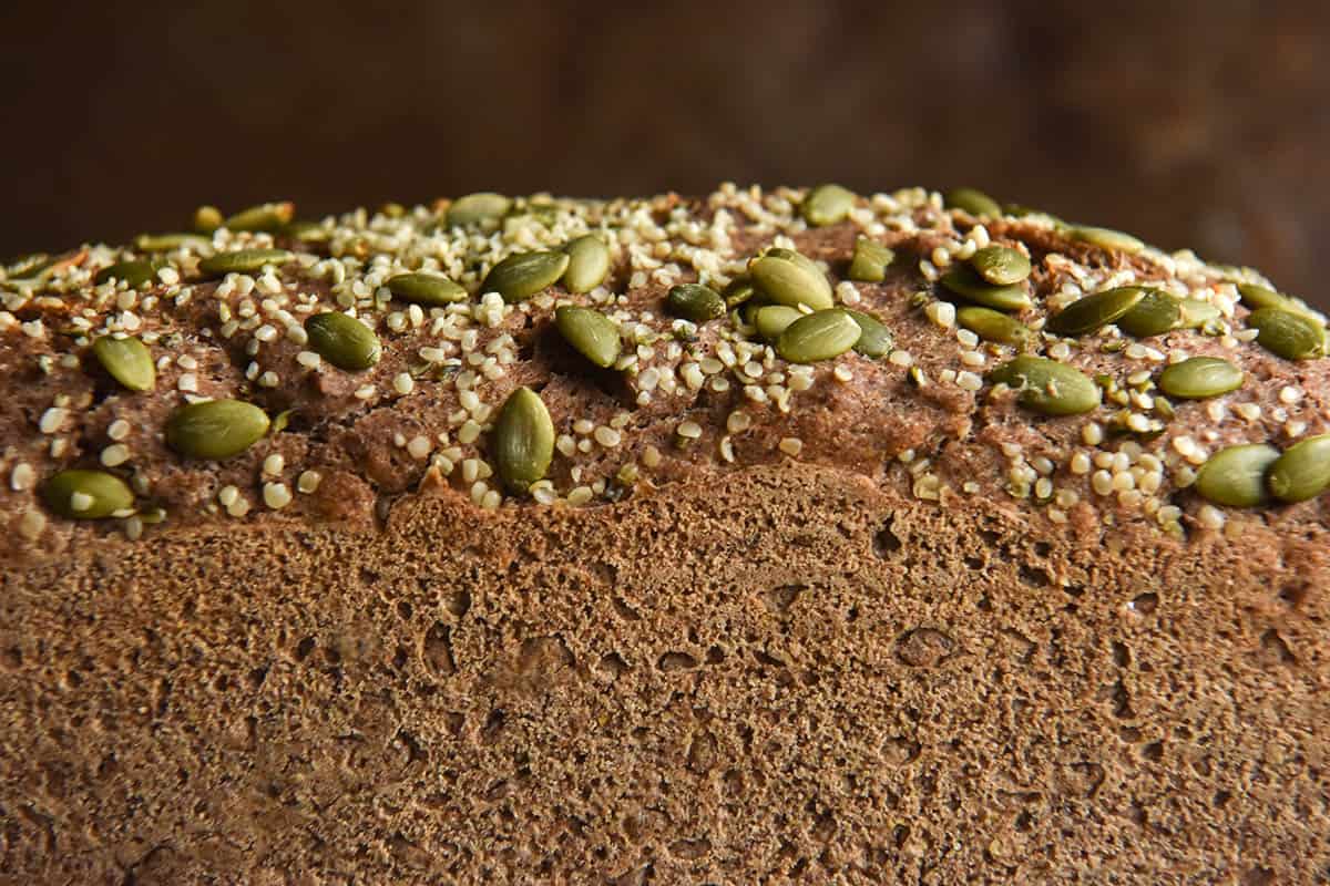 A macro side on image of a gluten free buckwheat bread loaf topped with with seeds. The loaf stands against an auburn coloured steel backdrop. 