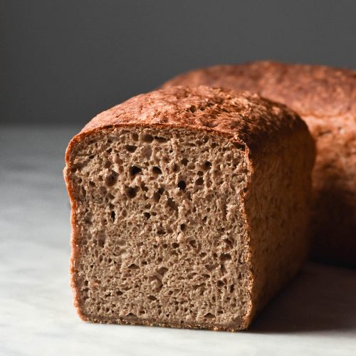 A side on view of a loaf of gluten free buckwheat bread on a white marble table. The loaf has been sliced and faces the camera, revealing the fluffy inner crumb. A second loaf sits off on angle behind the loaf.