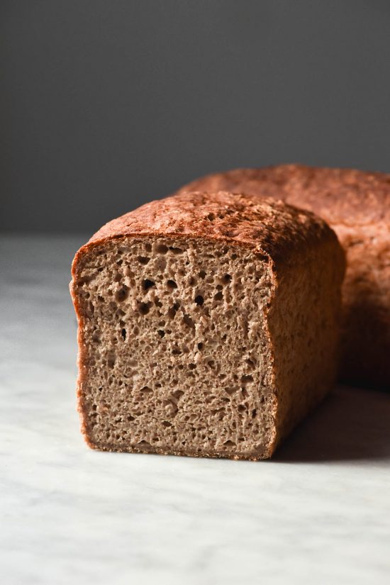 A side on view of a loaf of gluten free buckwheat bread on a white marble table. The loaf has been sliced and faces the camera, revealing the fluffy inner crumb. A second loaf sits off on angle behind the loaf.